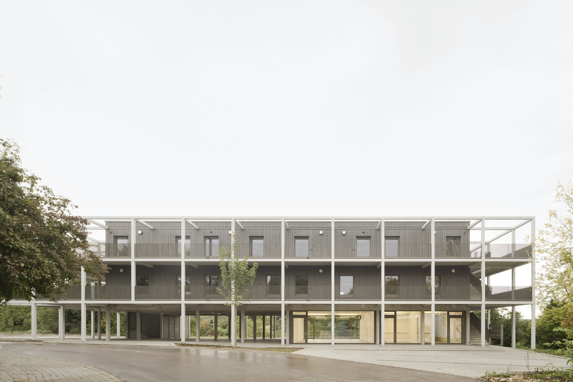 Front elevation showing the repeating module facade with white structural frame and concrete columns on a paved plaza