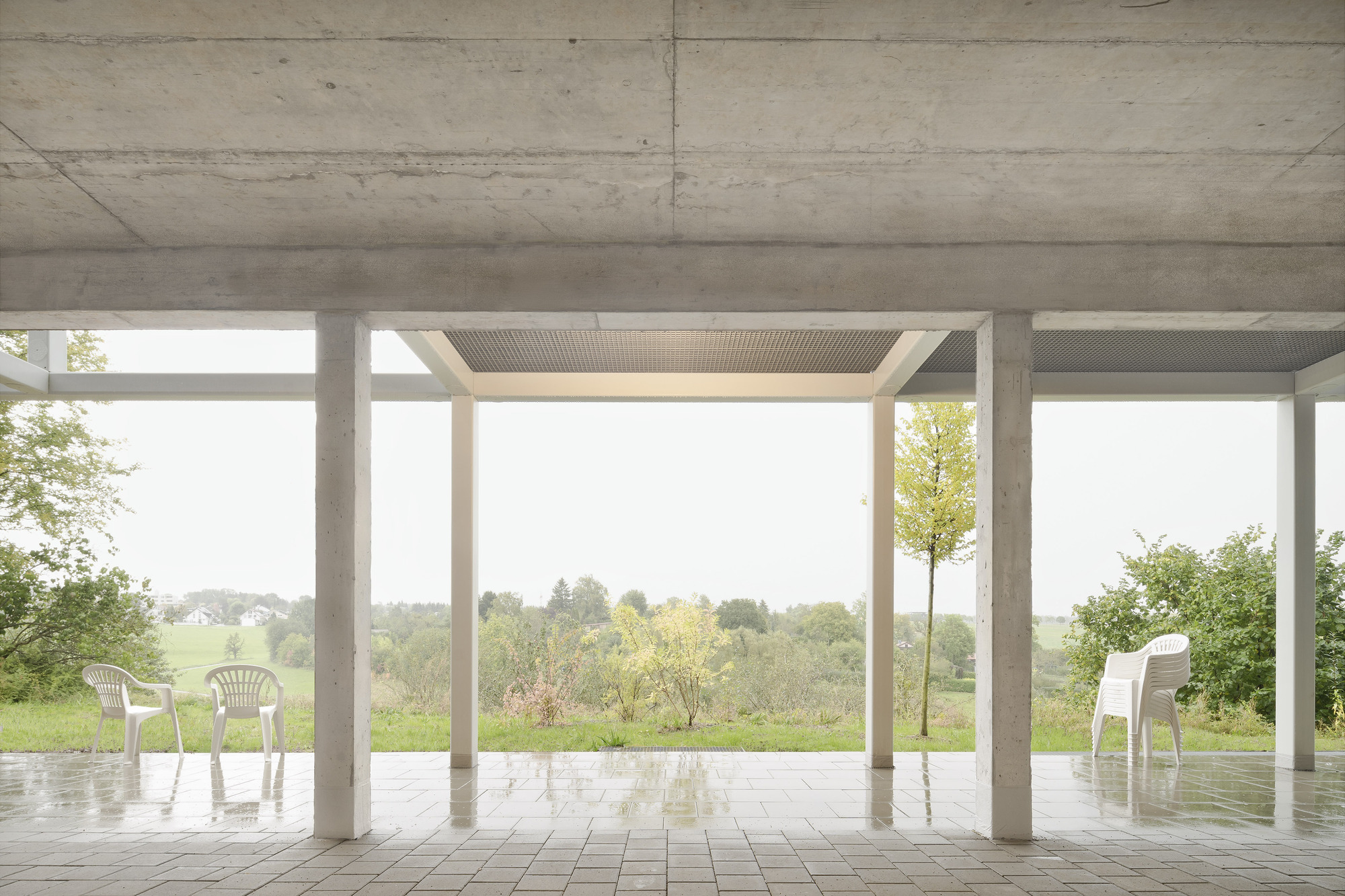 Covered ground-level terrace with exposed concrete ceiling and white plastic chairs overlooking fields and trees