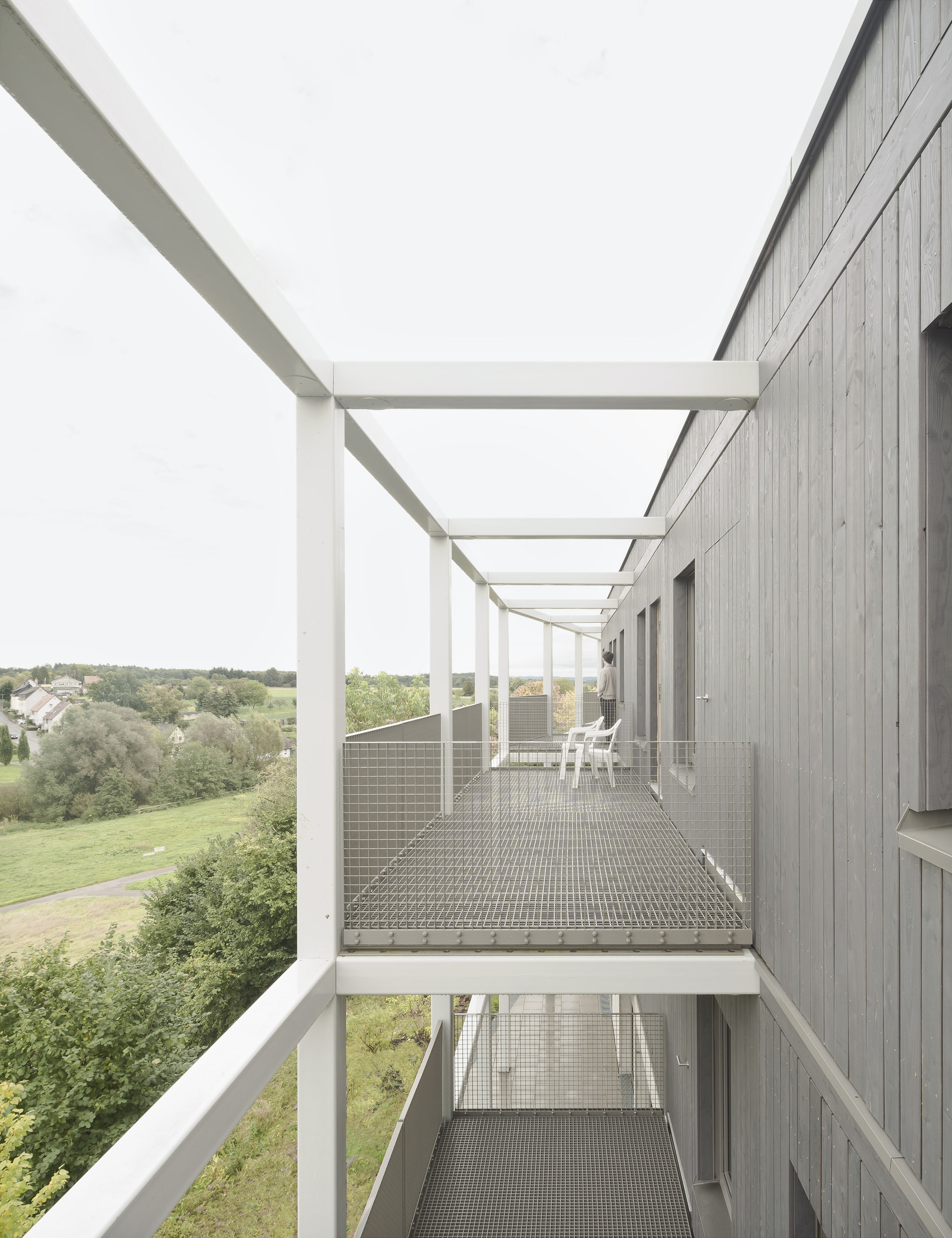 Exterior walkway with steel grating deck and white frame pergola overlooking a landscape of fields and trees