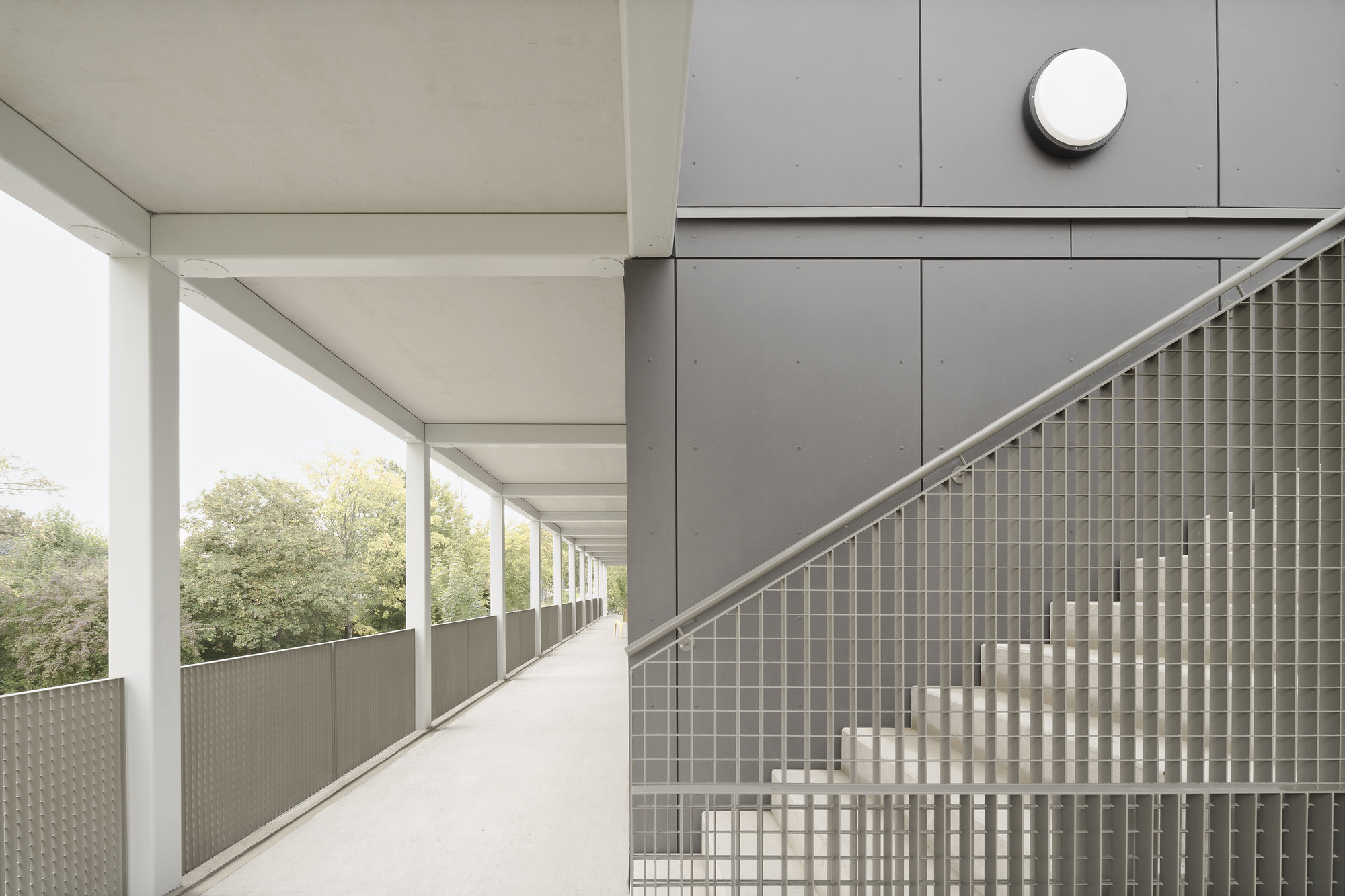 Exterior corridor with steel balustrade of woven metal mesh and gray panel cladding under overcast sky
