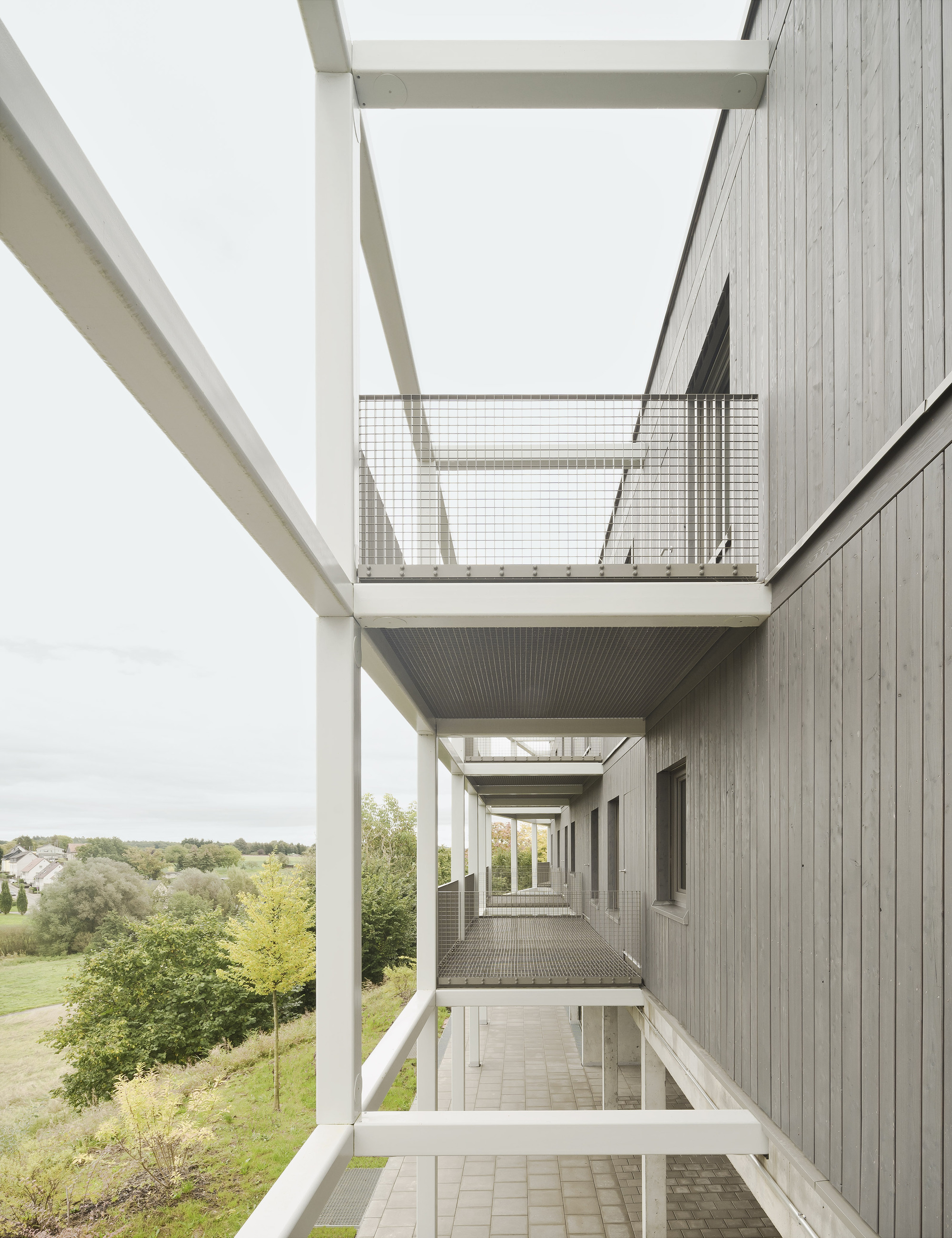 Three-story circulation void with white beams, wire mesh balconies and vertical timber cladding at dusk