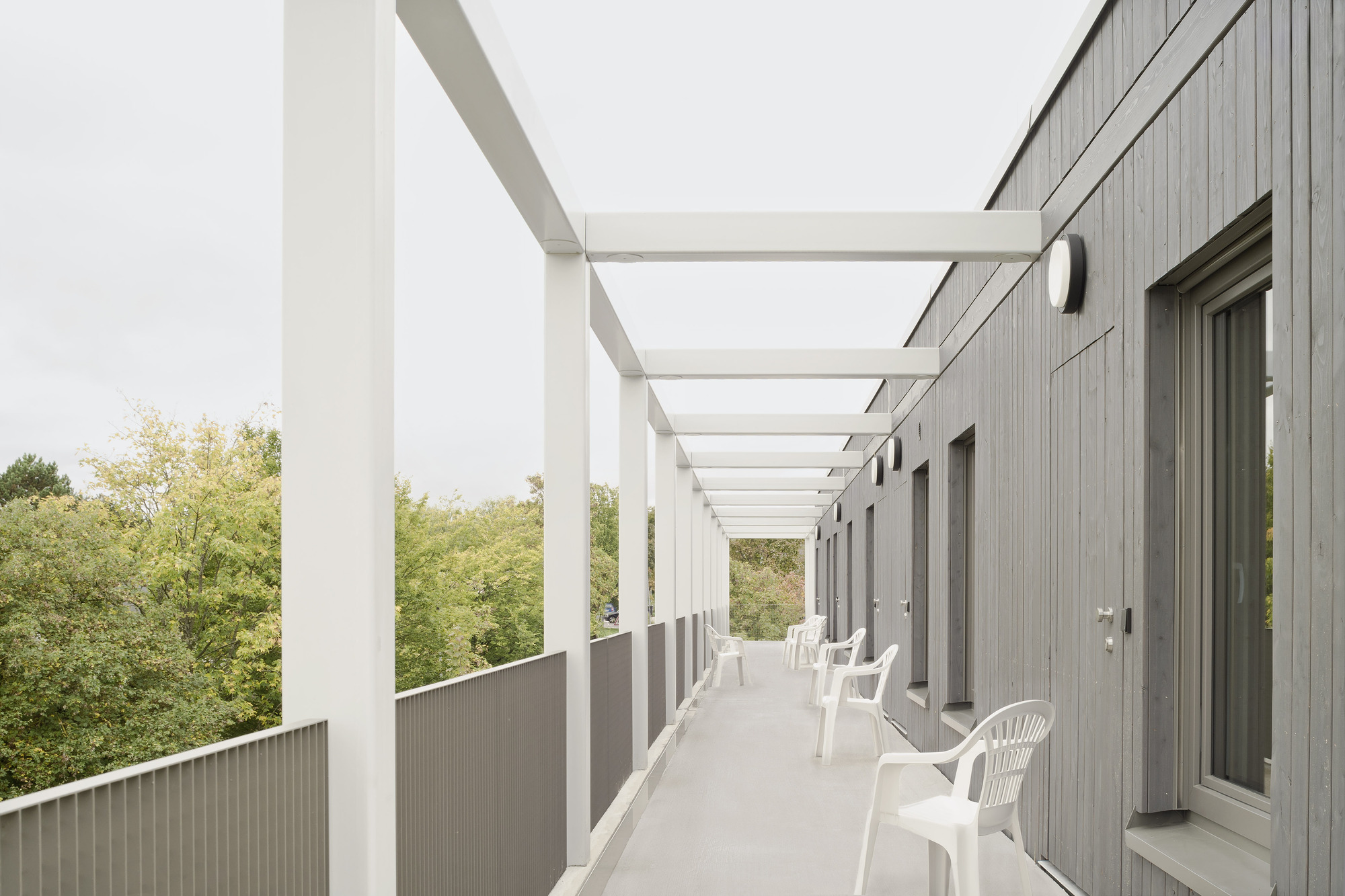 Exterior walkway with white steel pergola, grey cladding and white plastic chairs overlooking green trees