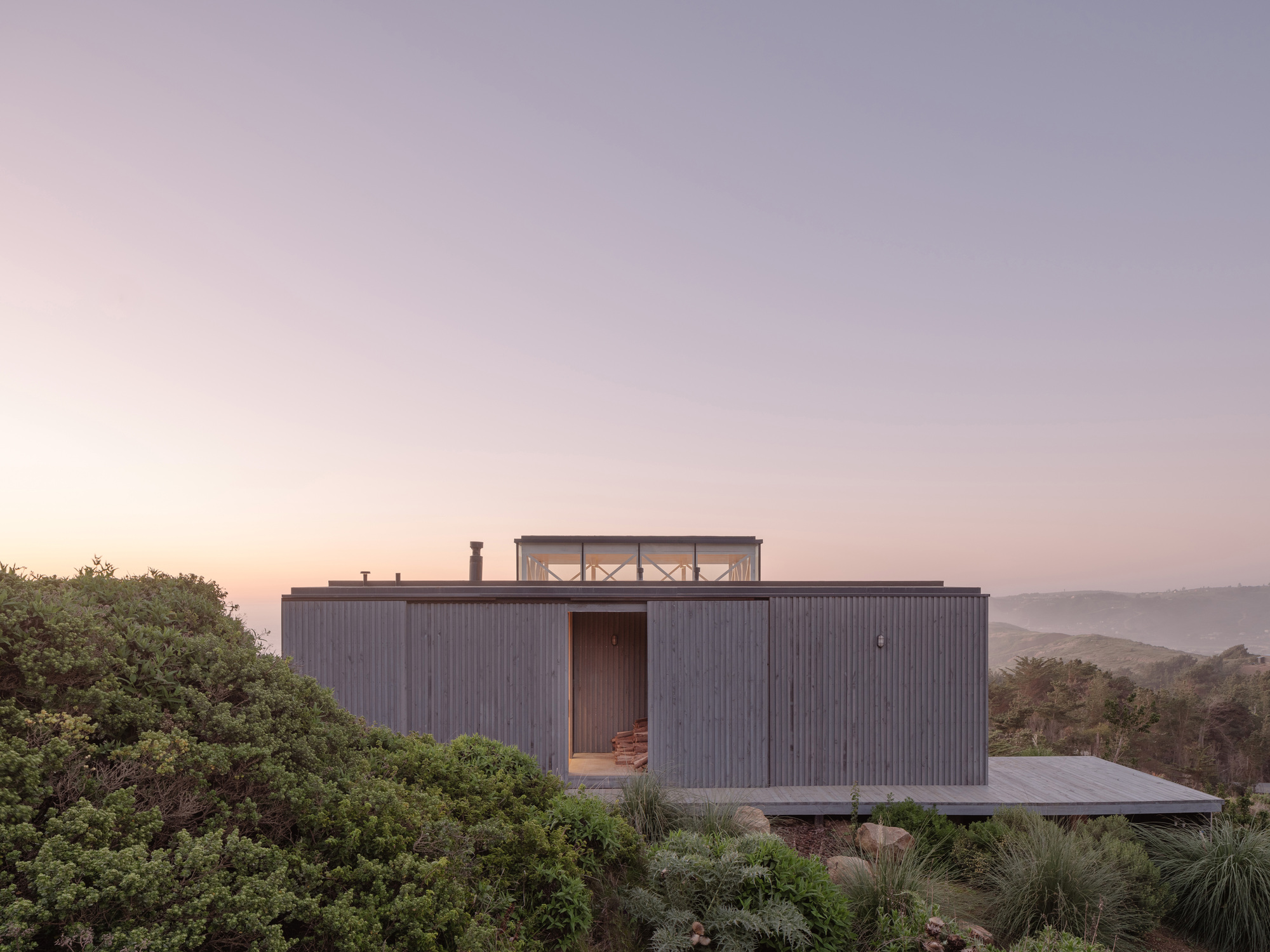 Vertically ribbed timber-clad volume with rooftop terrace overlooking coastal hills at dusk