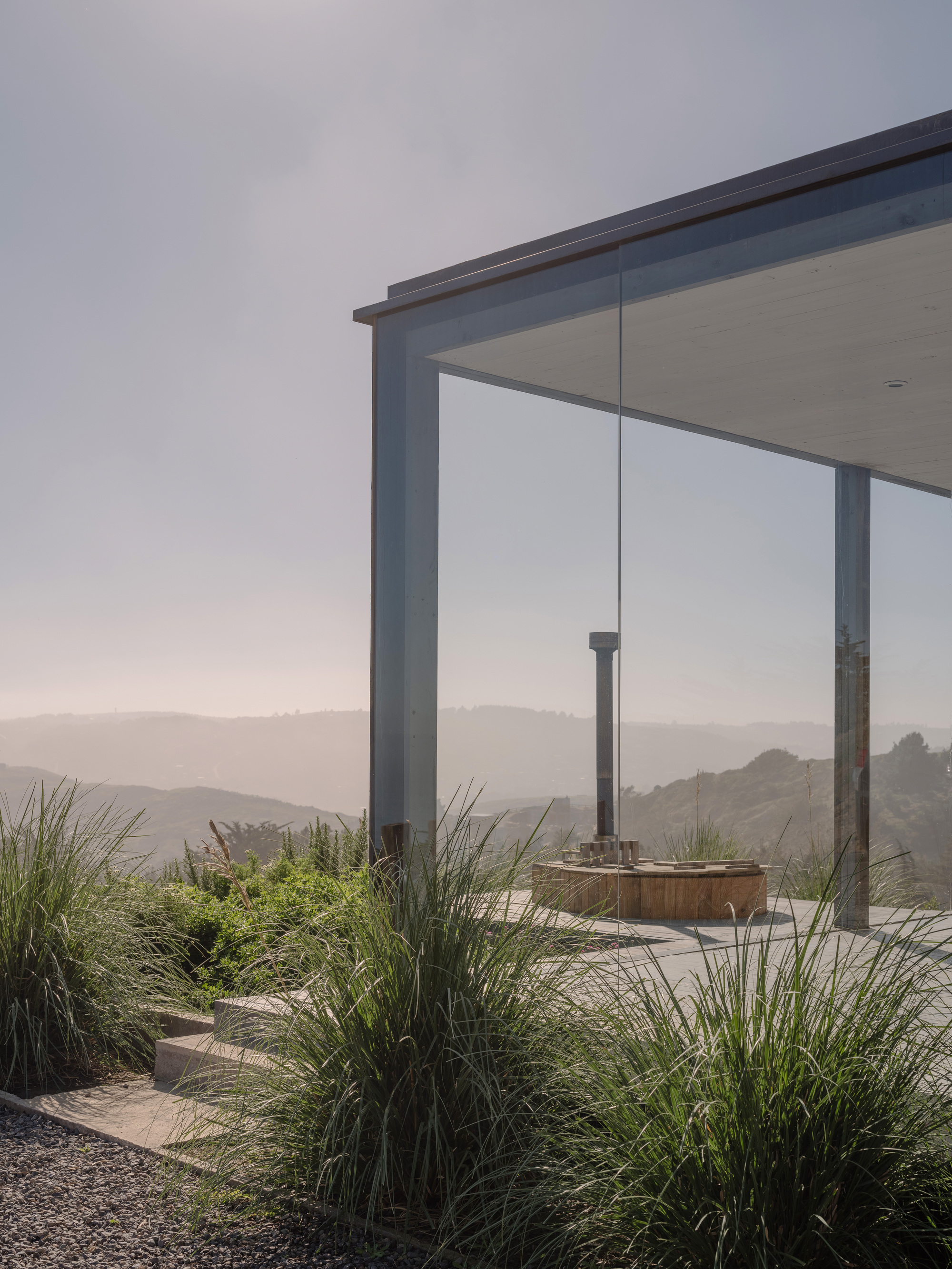Corner detail of glass walls meeting steel frame with hot tub and grasses in morning mist