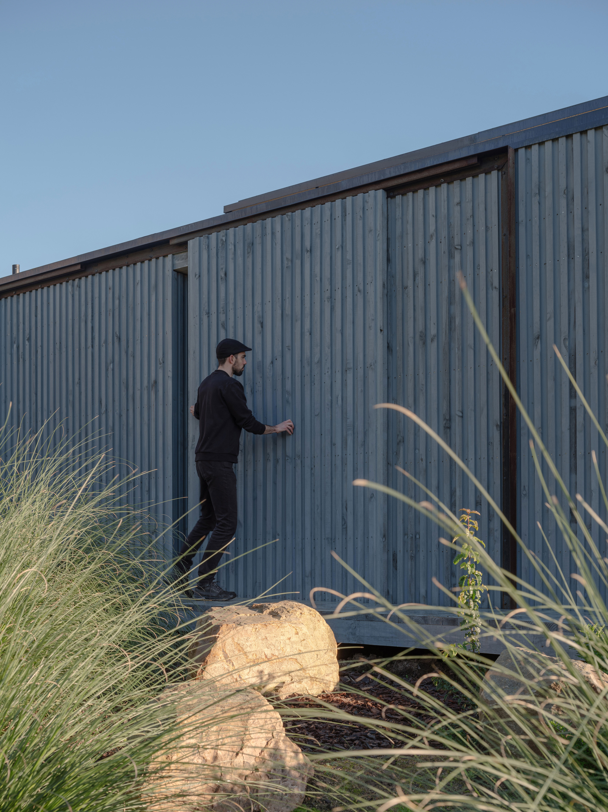 Figure opening sliding door on the corrugated timber facade framed by grasses and boulders