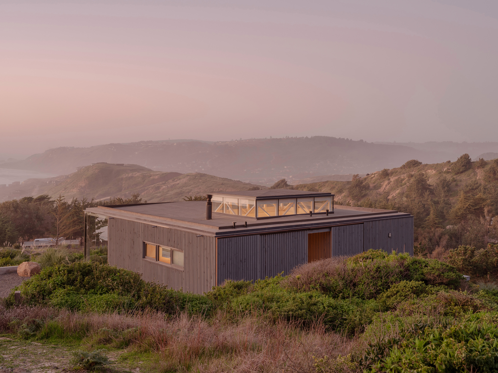 Corrugated metal residence with glazed rooftop volume nestled into hillside scrub at sunset