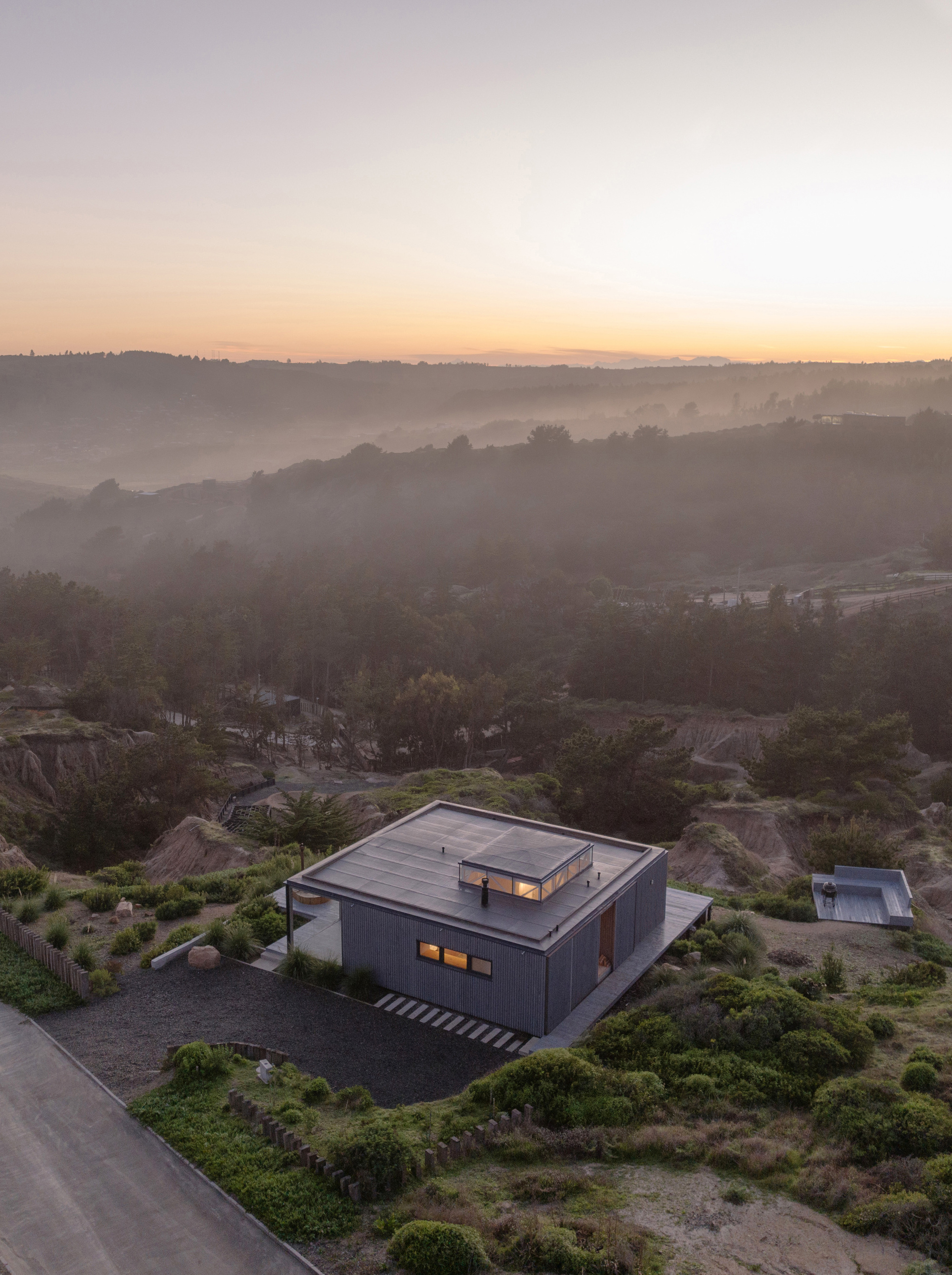 Aerial view of the corrugated metal-clad volume nestled among rocky hillside vegetation at dusk with mist