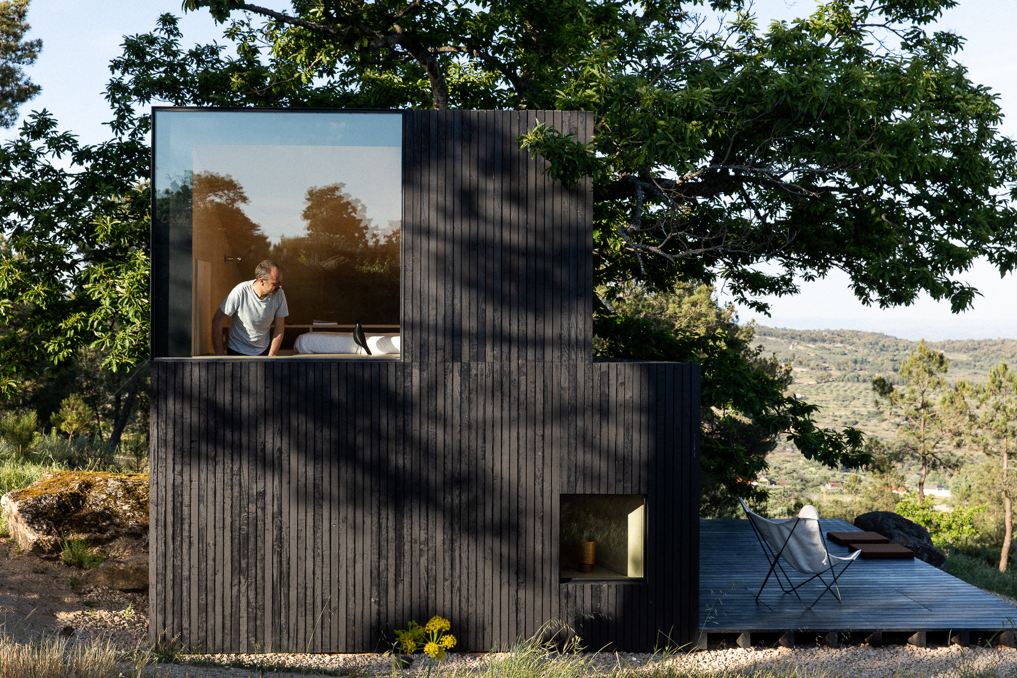 Charred timber volume with large window framing an occupant, nestled among oak trees on a hillside