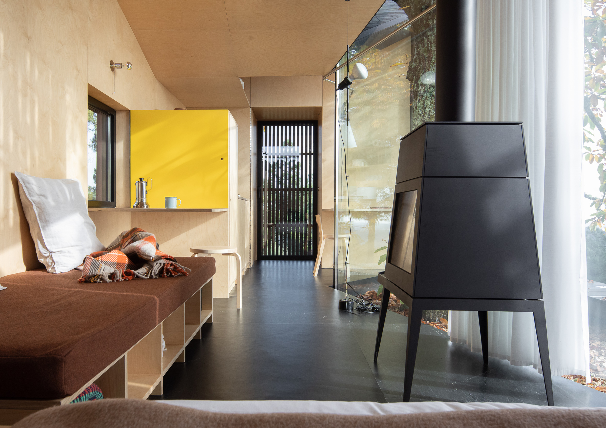 Interior living space with plywood ceiling, black metal stove, yellow cabinet, and glass wall