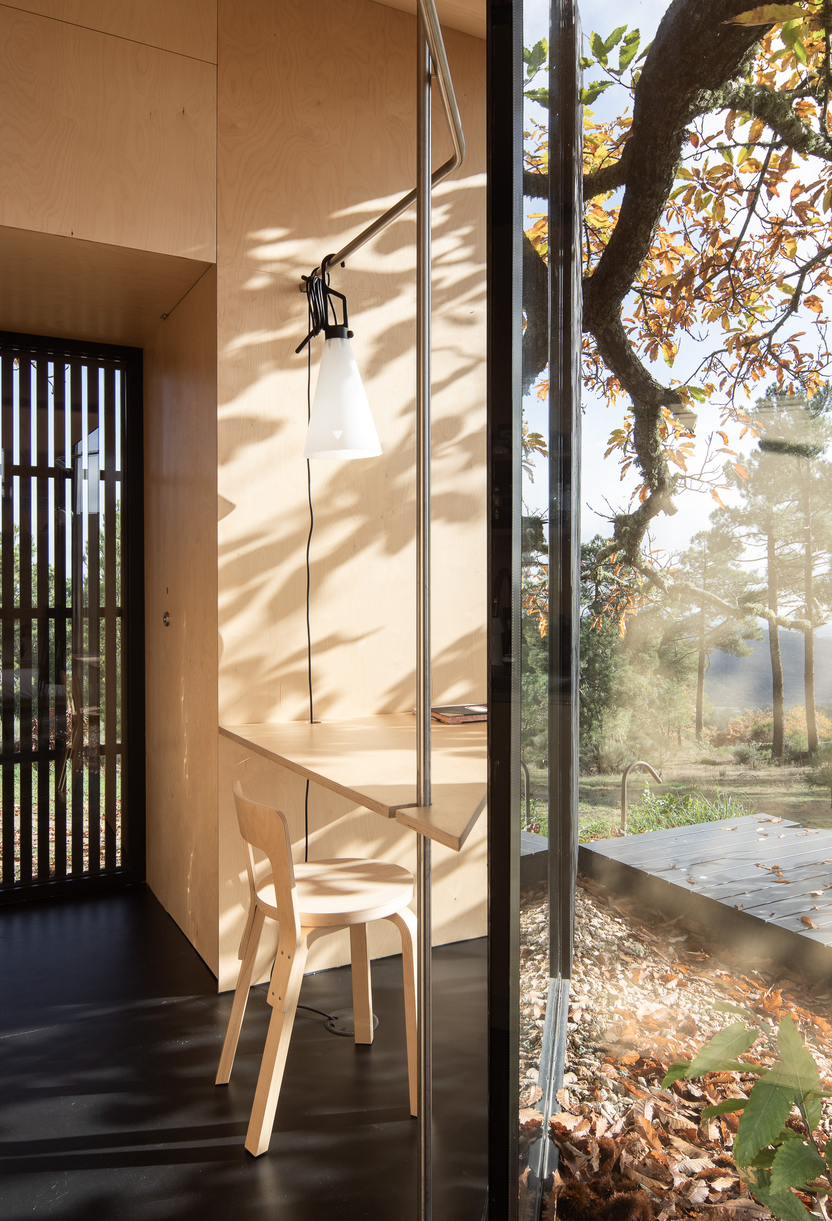 Plywood desk and chair beneath a window casting dappled sunlight through tree branches