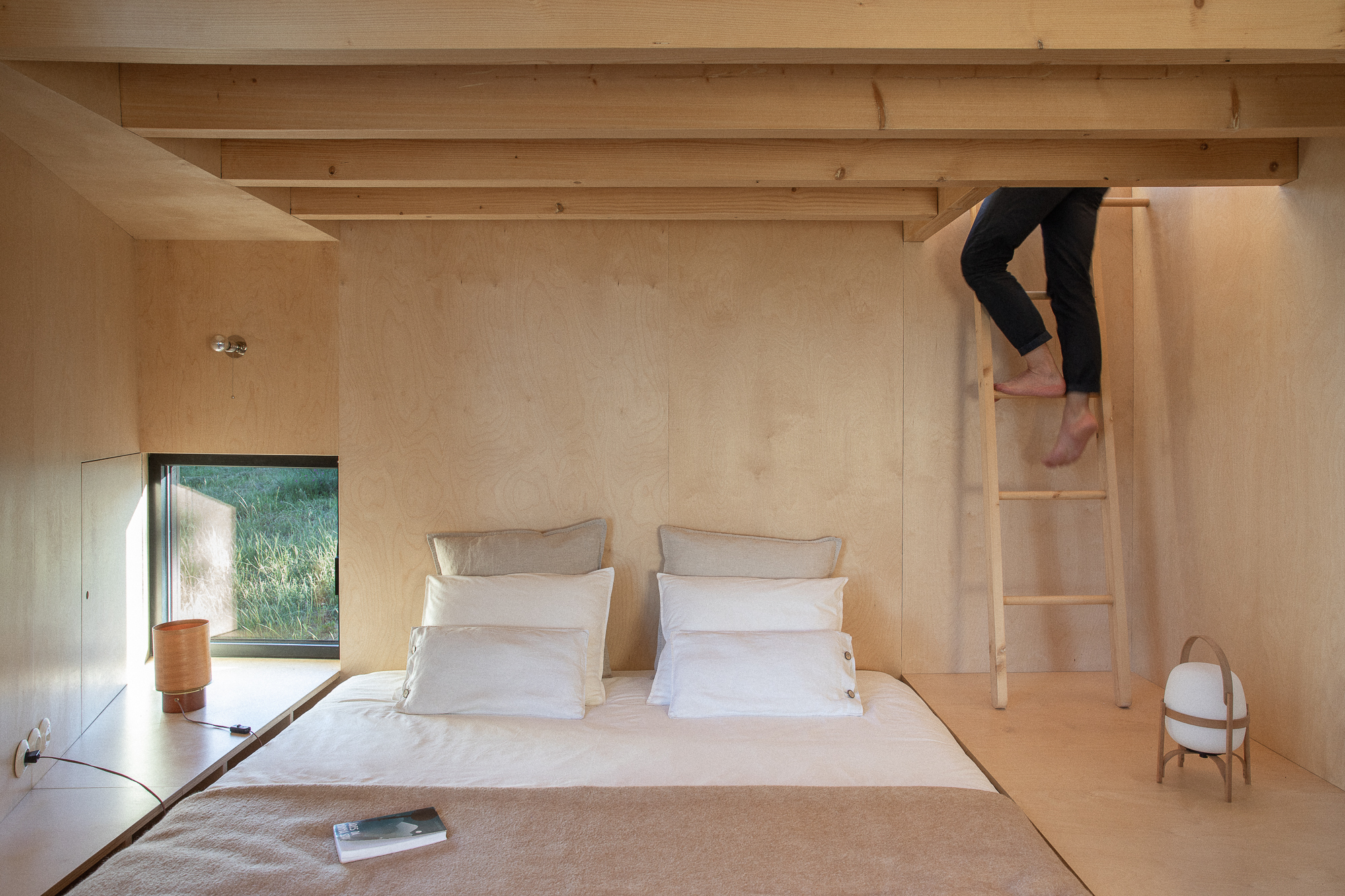 Plywood bedroom with exposed timber beams and wooden ladder where a person climbs toward an upper level