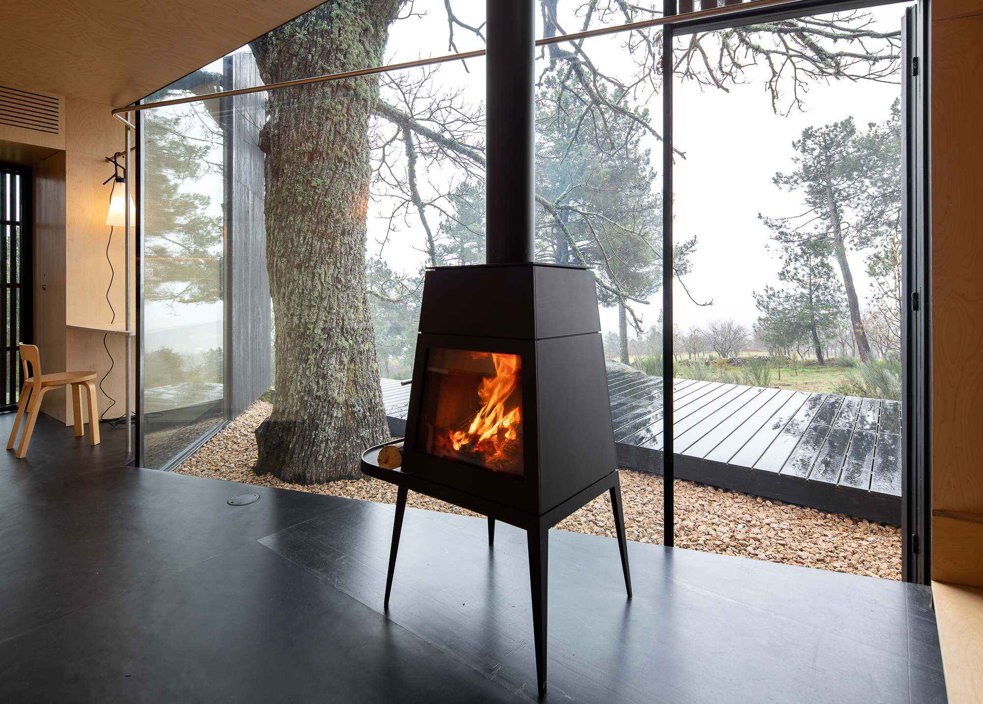 Black metal wood stove with flue pipe beside floor-to-ceiling glazing overlooking a misty pine forest