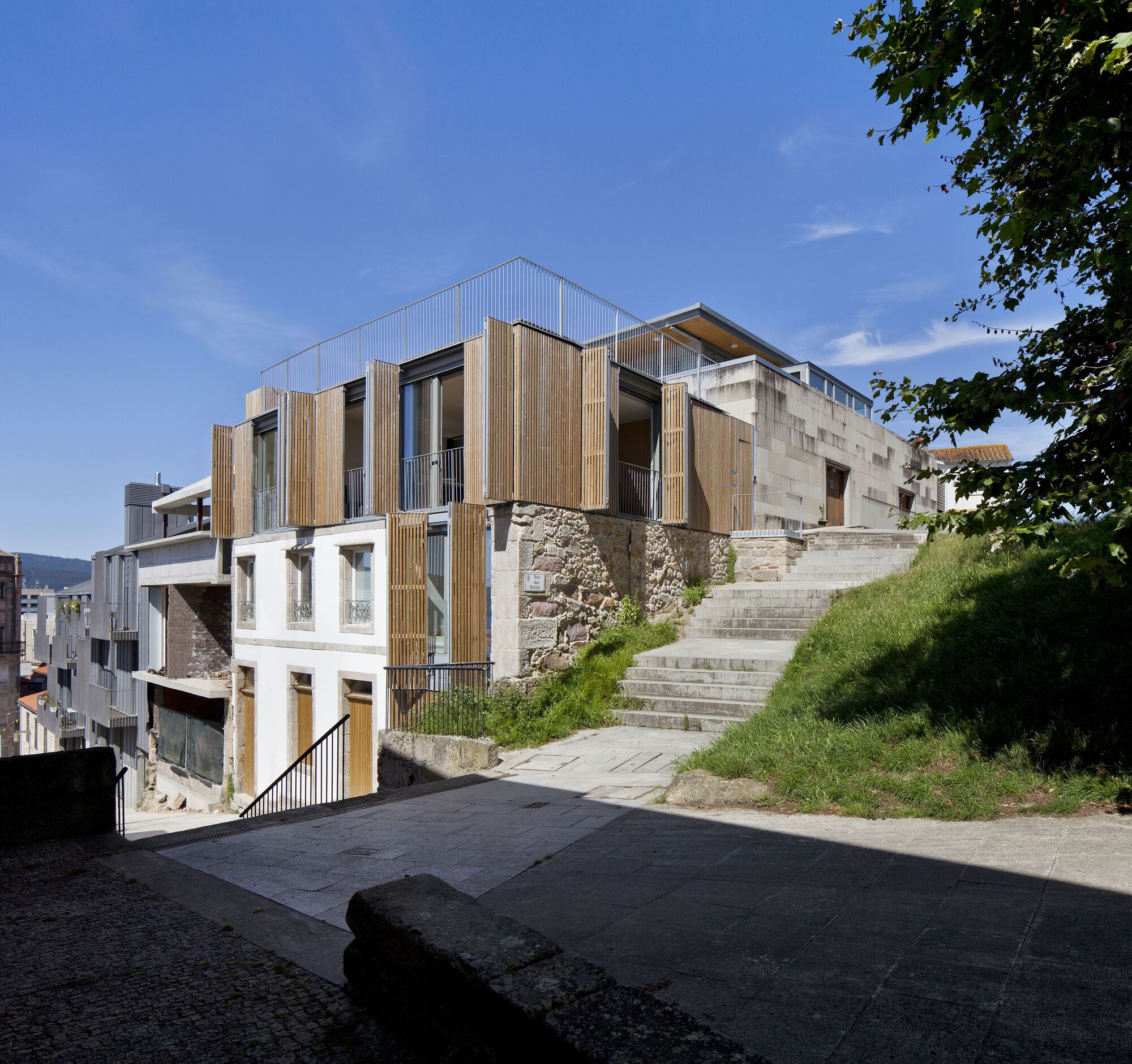 Street view of timber-clad volumes and concrete facades rising from a stone retaining wall with grassy slope