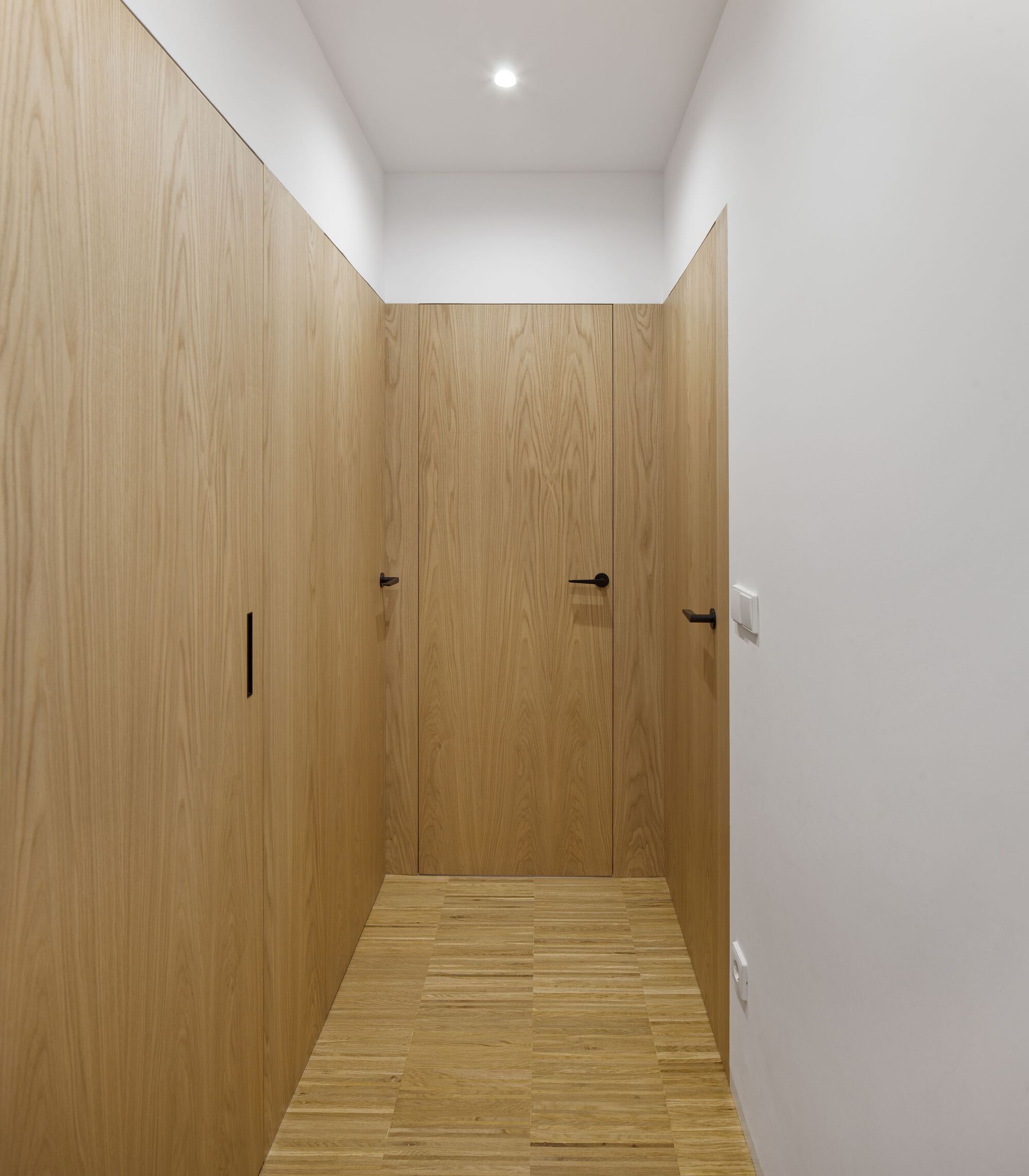 Corridor lined with flush oak veneer panels concealing doors and storage under recessed ceiling light
