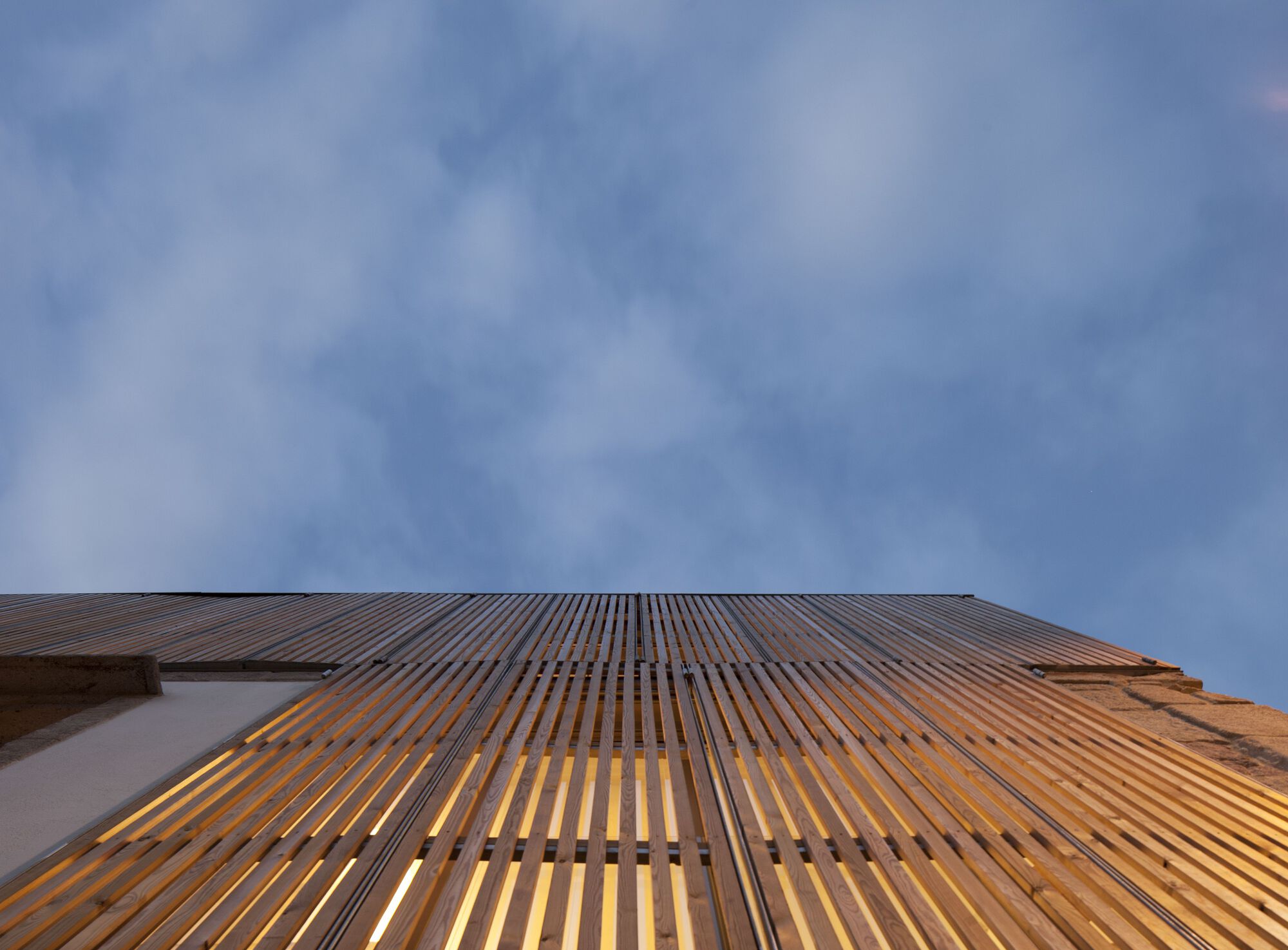 Timber slat screen facade illuminated from within at dusk beneath stormy clouds