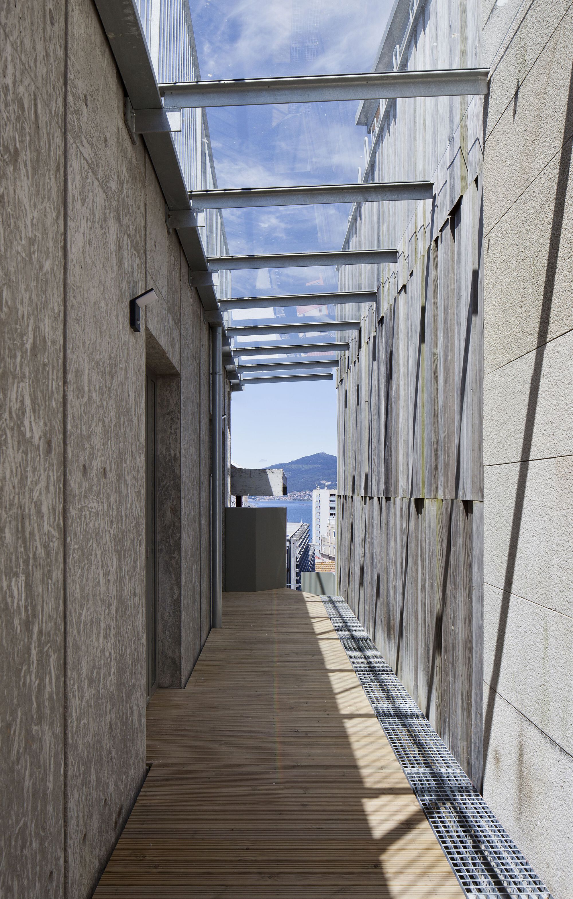 Timber deck corridor with overhead glass canopy between wood-clad and concrete walls framing a harbor view