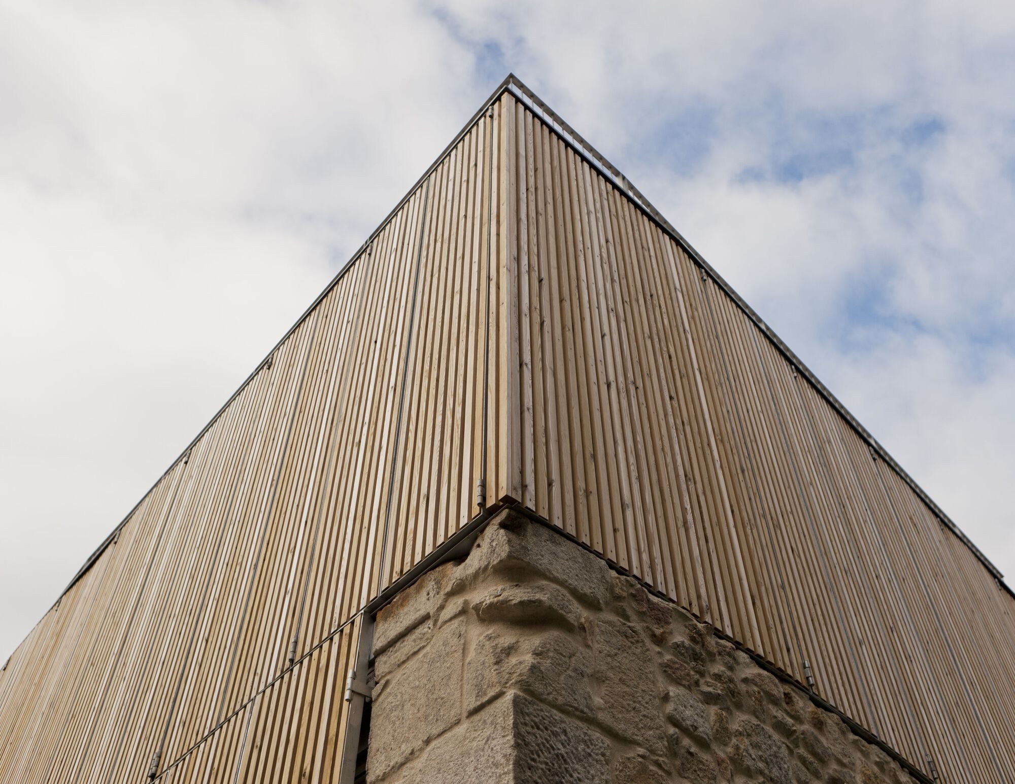 Upward view of the vertical timber cladding meeting an existing stone corner under cloudy skies