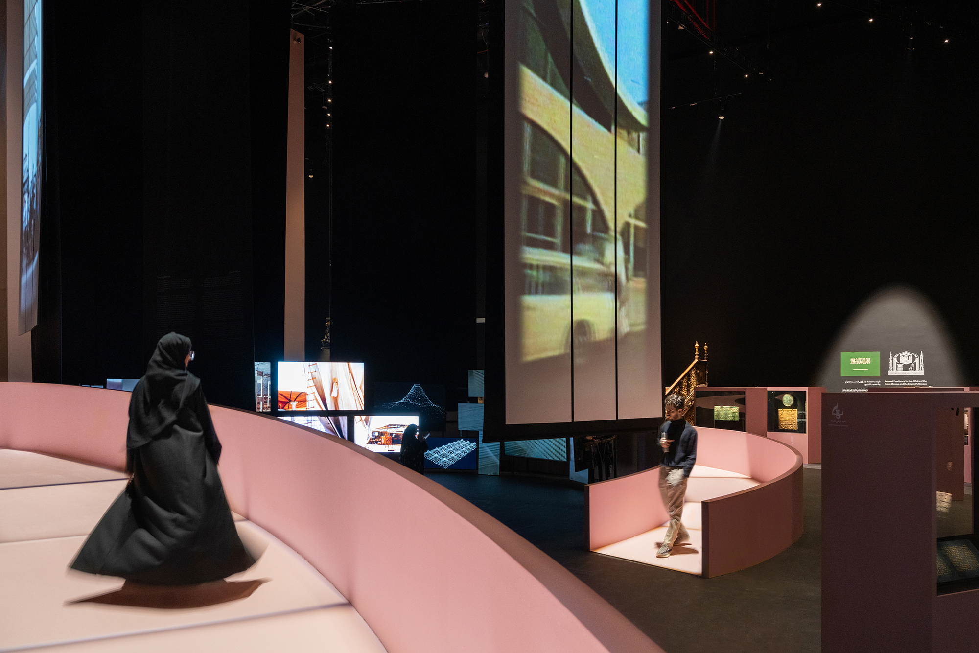Curved pink platform with visitors and interactive displays beneath projected video on gallery walls