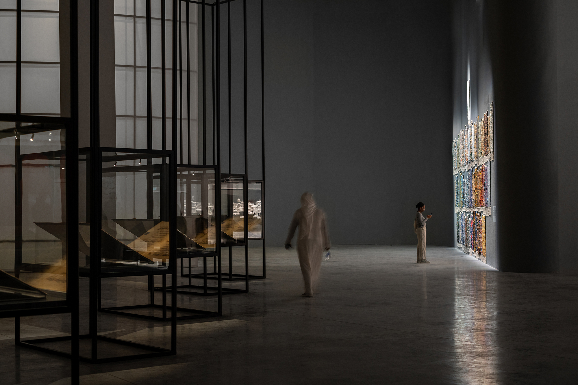Gallery hall with black steel frame shelving and visitors near illuminated textile hanging on wall