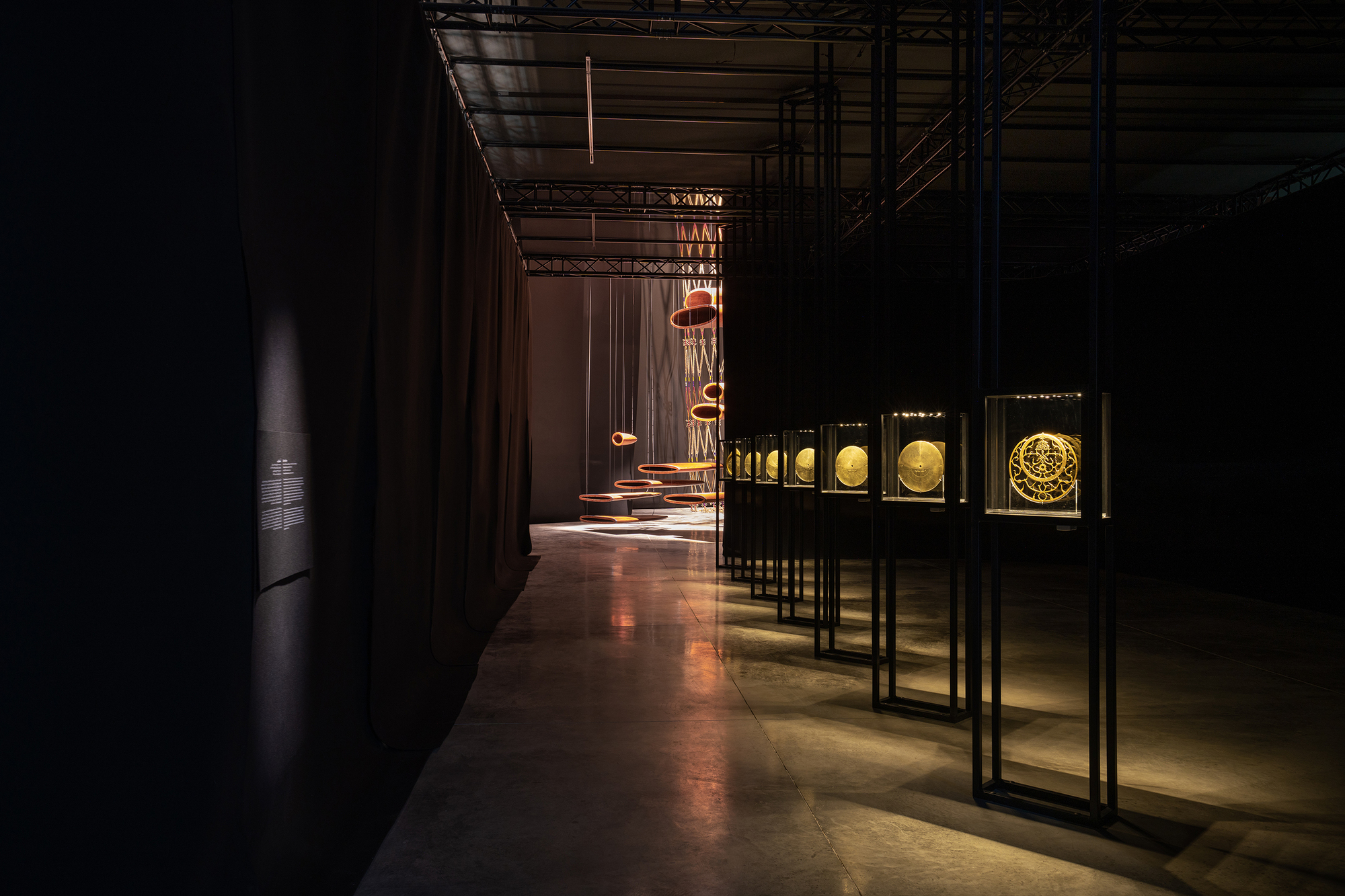Exhibition corridor with backlit artifact cases and suspended copper lighting elements under exposed ceiling infrastructure