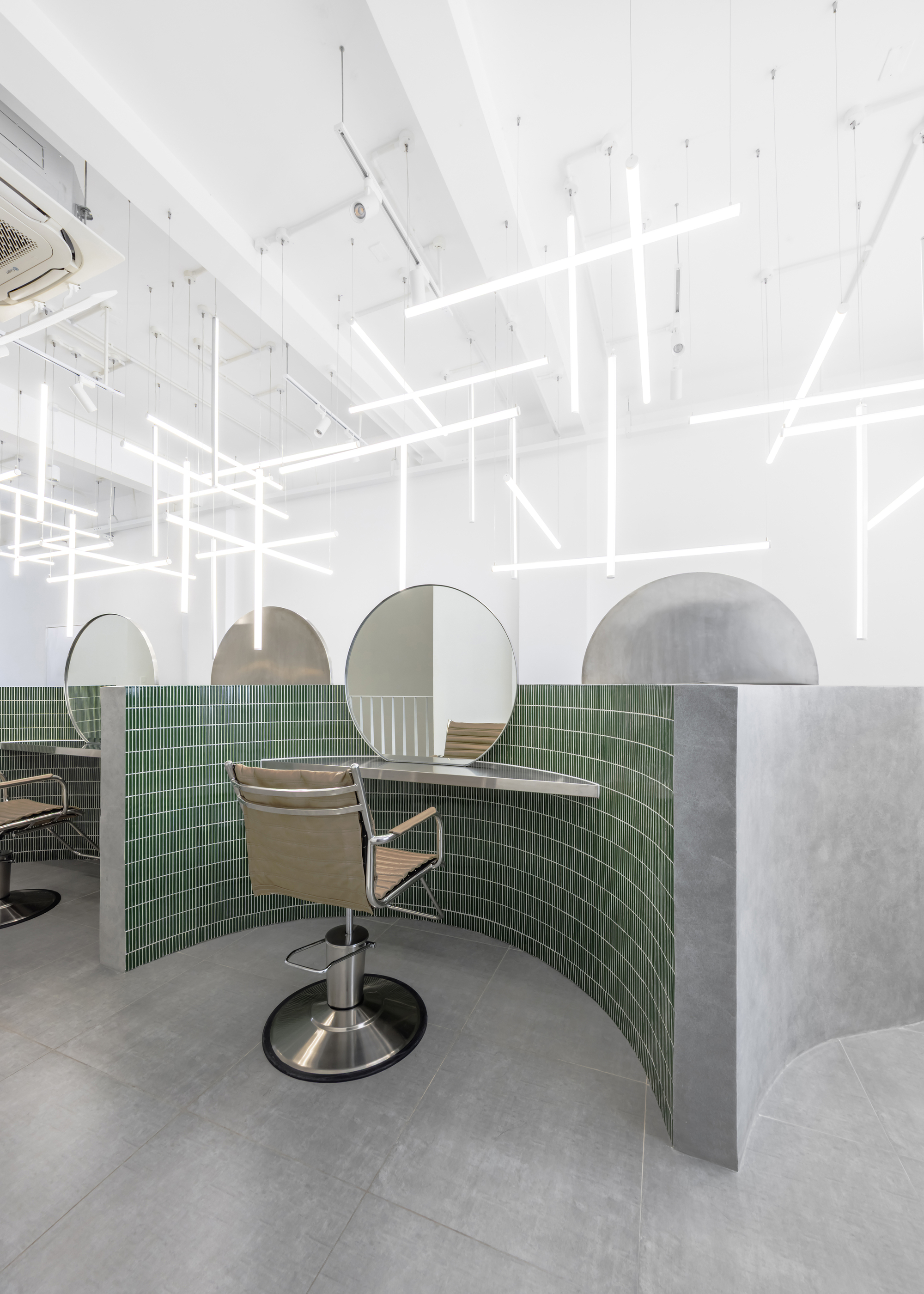 Salon station with curved green tile partition, circular mirror, and hydraulic chair under gridded ceiling lights