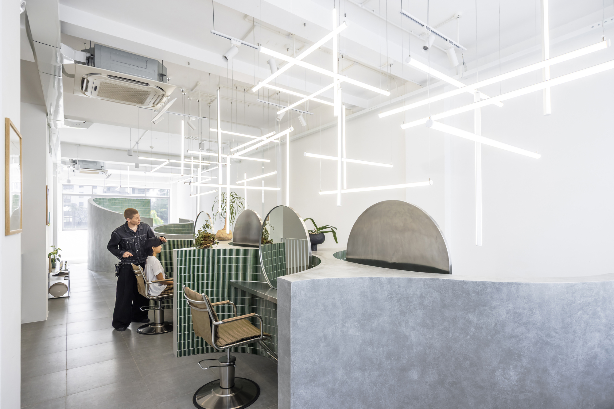White grid light fixture suspended over green tiled salon stations with chrome chairs and a stylist at work