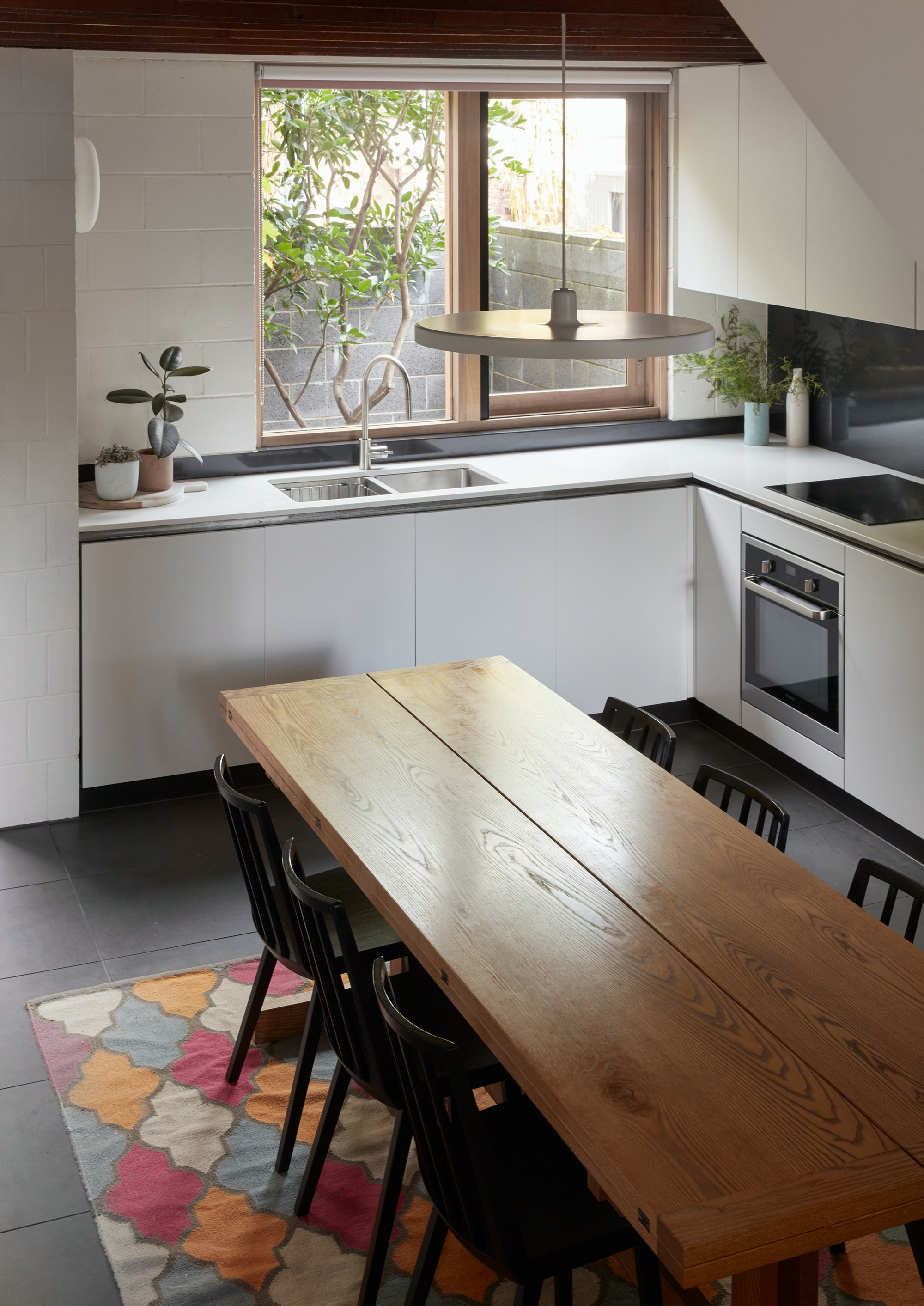 Kitchen with timber dining table, white cabinetry, and window view to courtyard garden and tree