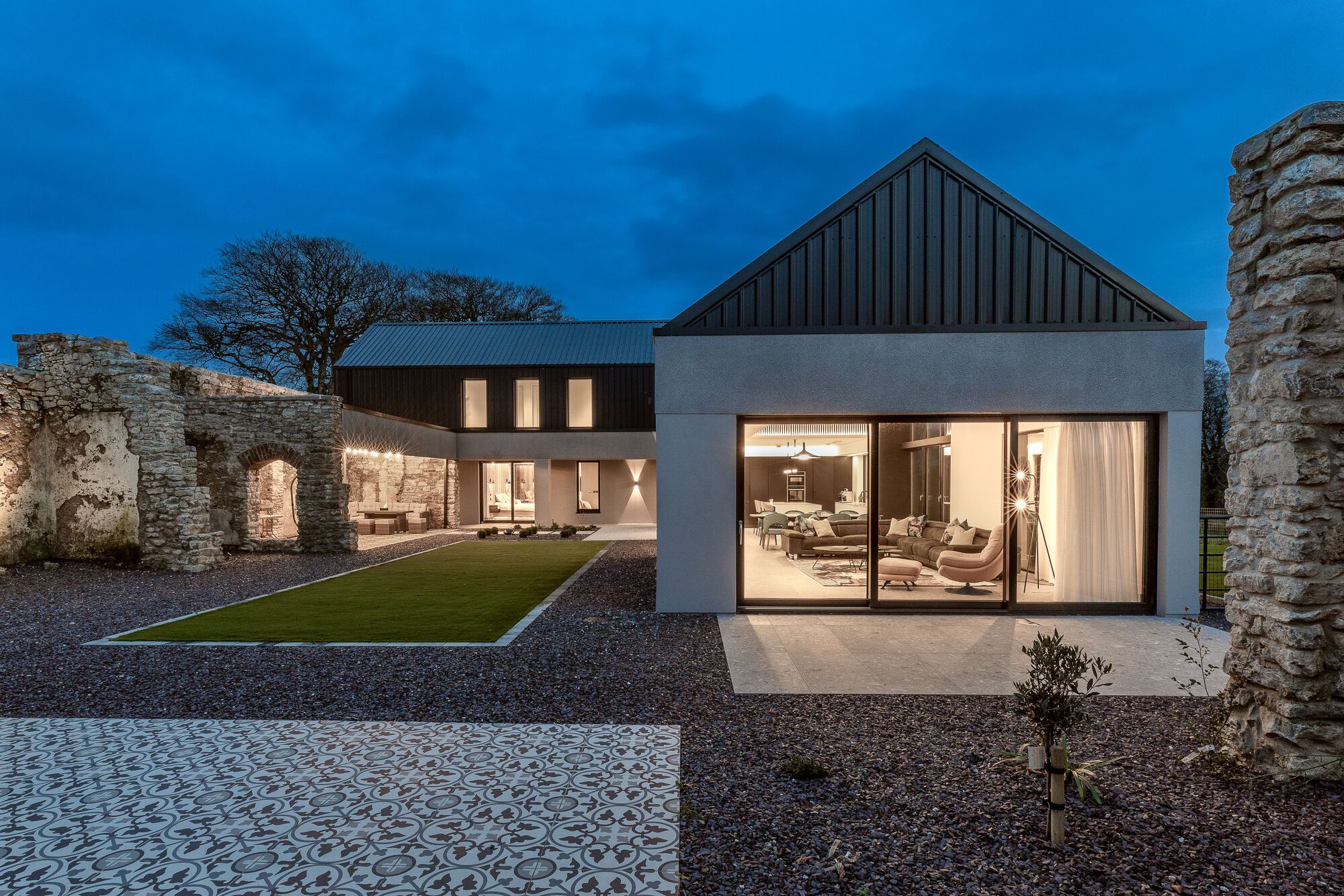 Glass living pavilion opening to the courtyard with metal roofed volumes and stone walls at dusk