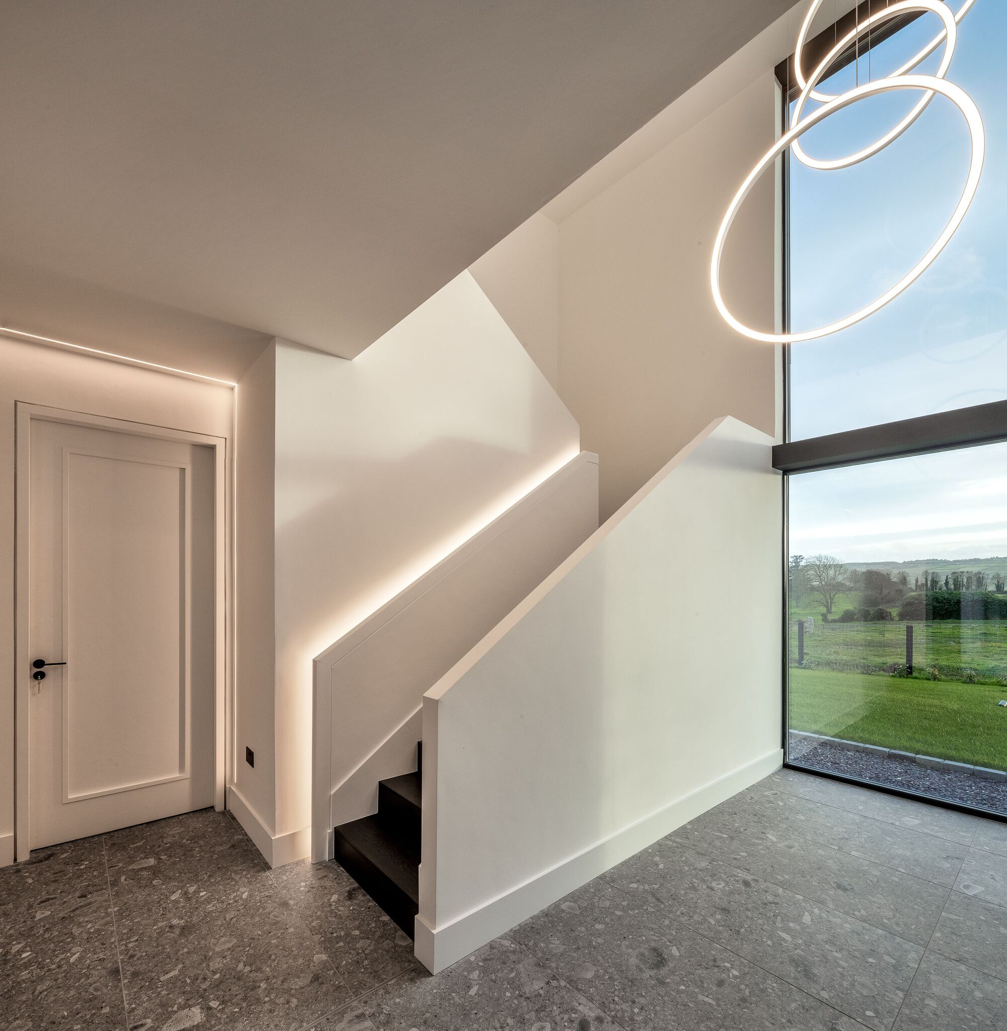 Entry hall showing illuminated stair balustrade and floor-to-ceiling glazing overlooking green fields