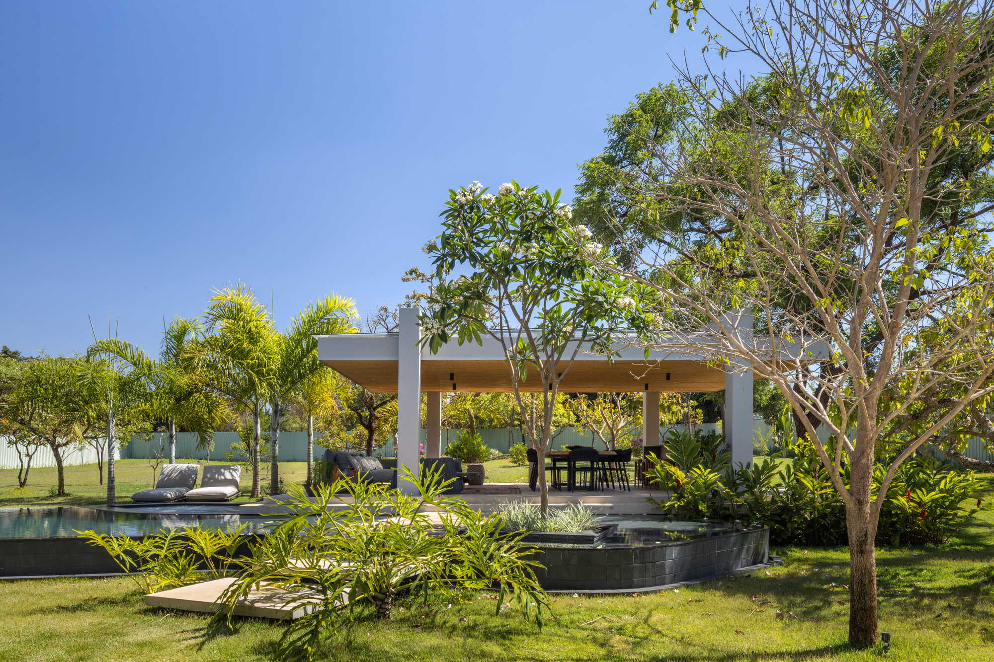 Open pavilion with flat roof surrounded by lawn and mature trees under a clear sky