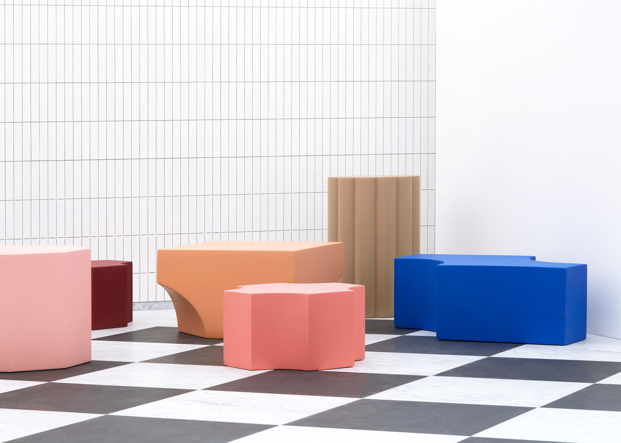 Colored seating blocks in pink, coral, and blue arranged on a chequered floor in bright sunlight
