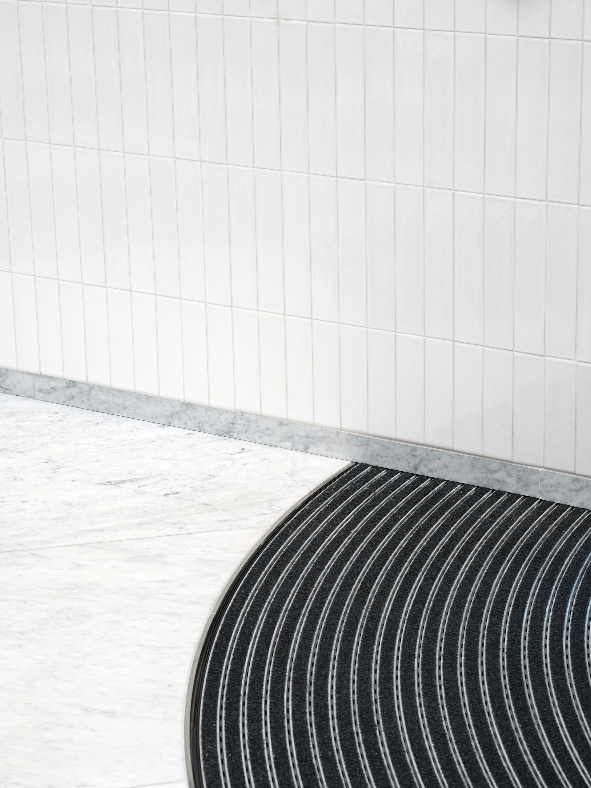 Curved black ribbed floor mat meeting white vertical tile wall with terrazzo base trim