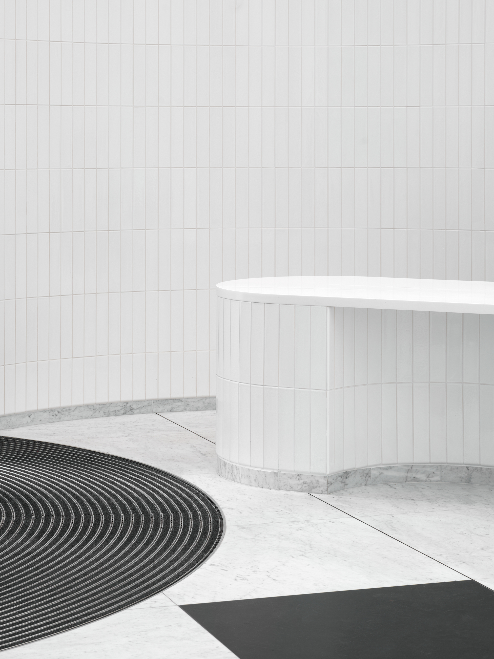 White cylindrical reception desk beside curved black floor mat on terrazzo in a tiled lobby