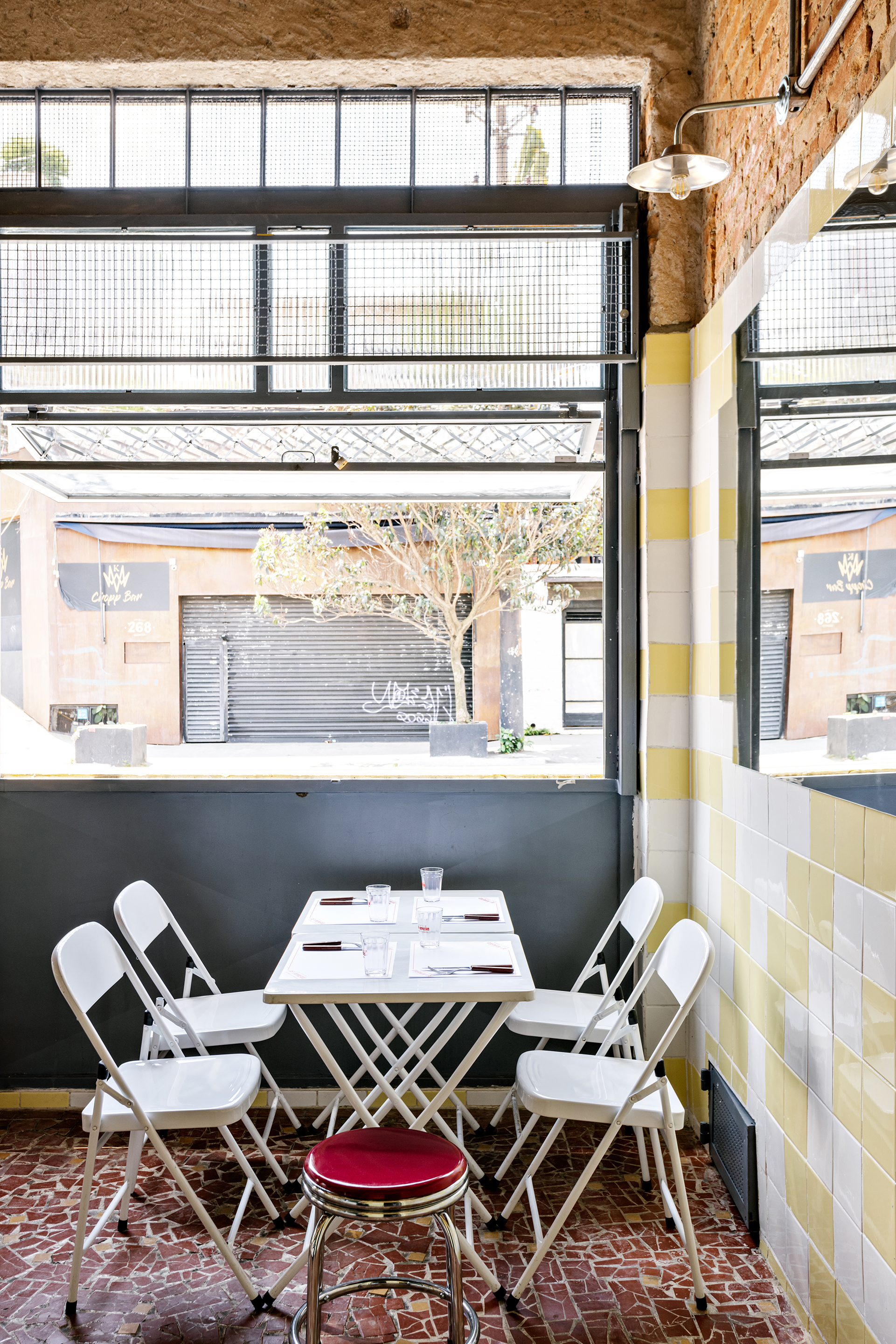 Dining table with white folding chairs positioned below a steel-framed window overlooking the street