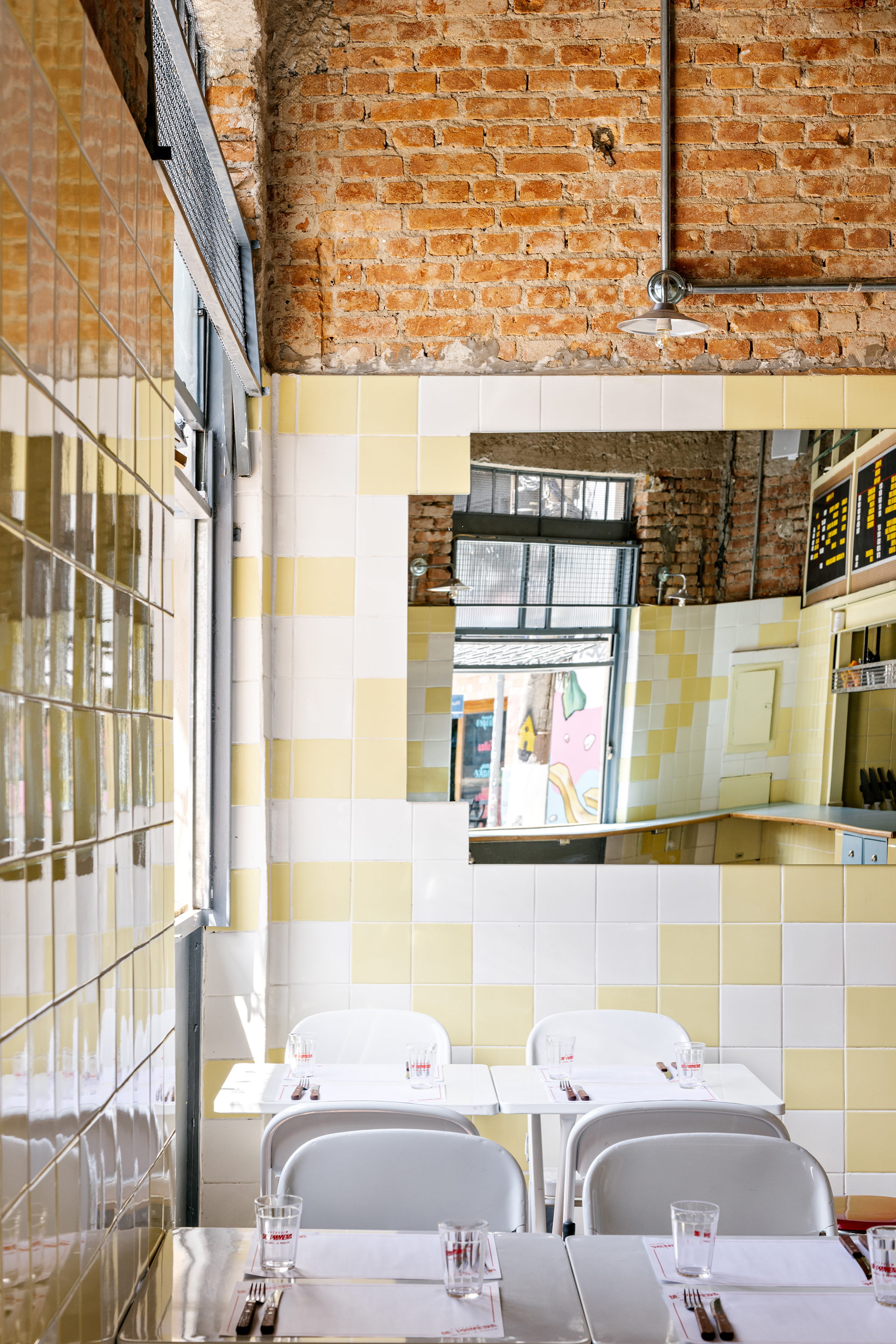 Dining tables beneath exposed brick and a mirrored wall reflecting yellow and white checkerboard tile surfaces