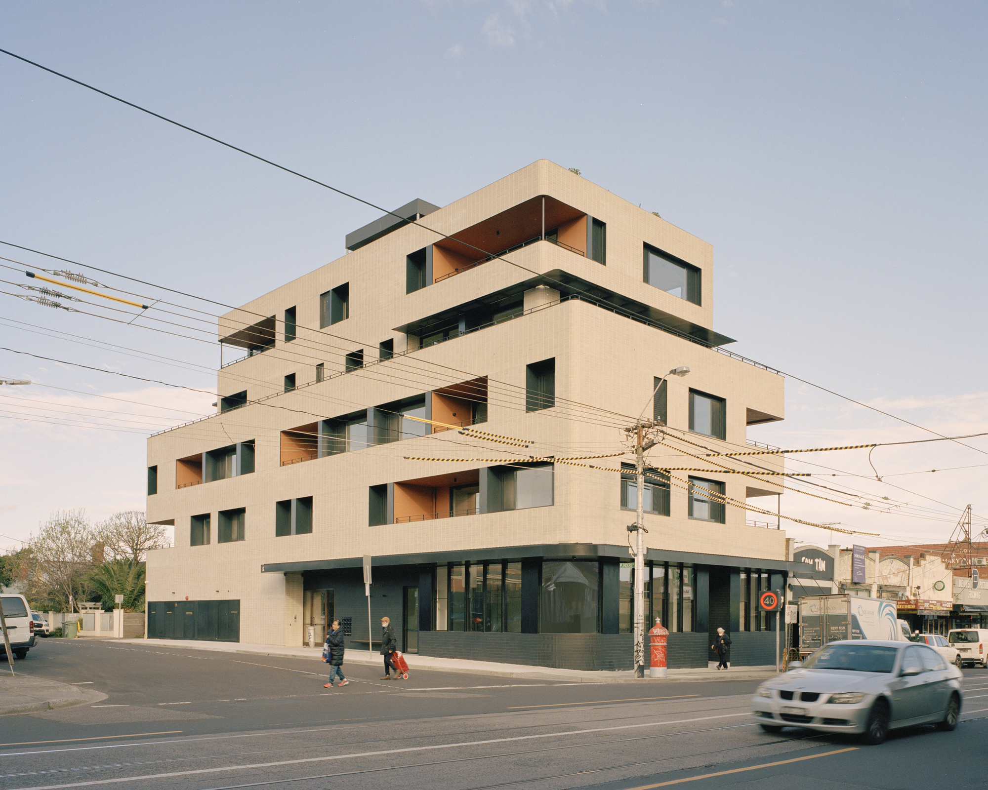 Corner view of the stacked residential volume with pale tile cladding and pedestrians crossing below power lines