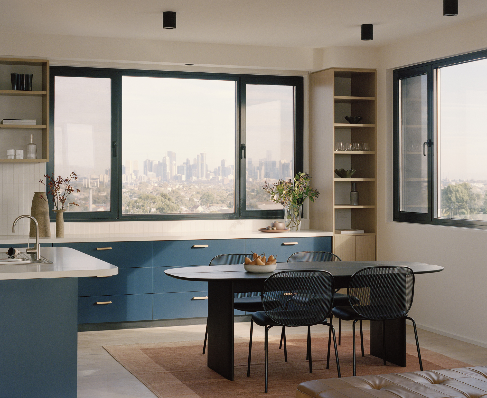 Open-plan kitchen and dining area with blue cabinetry and black-framed windows overlooking the city skyline