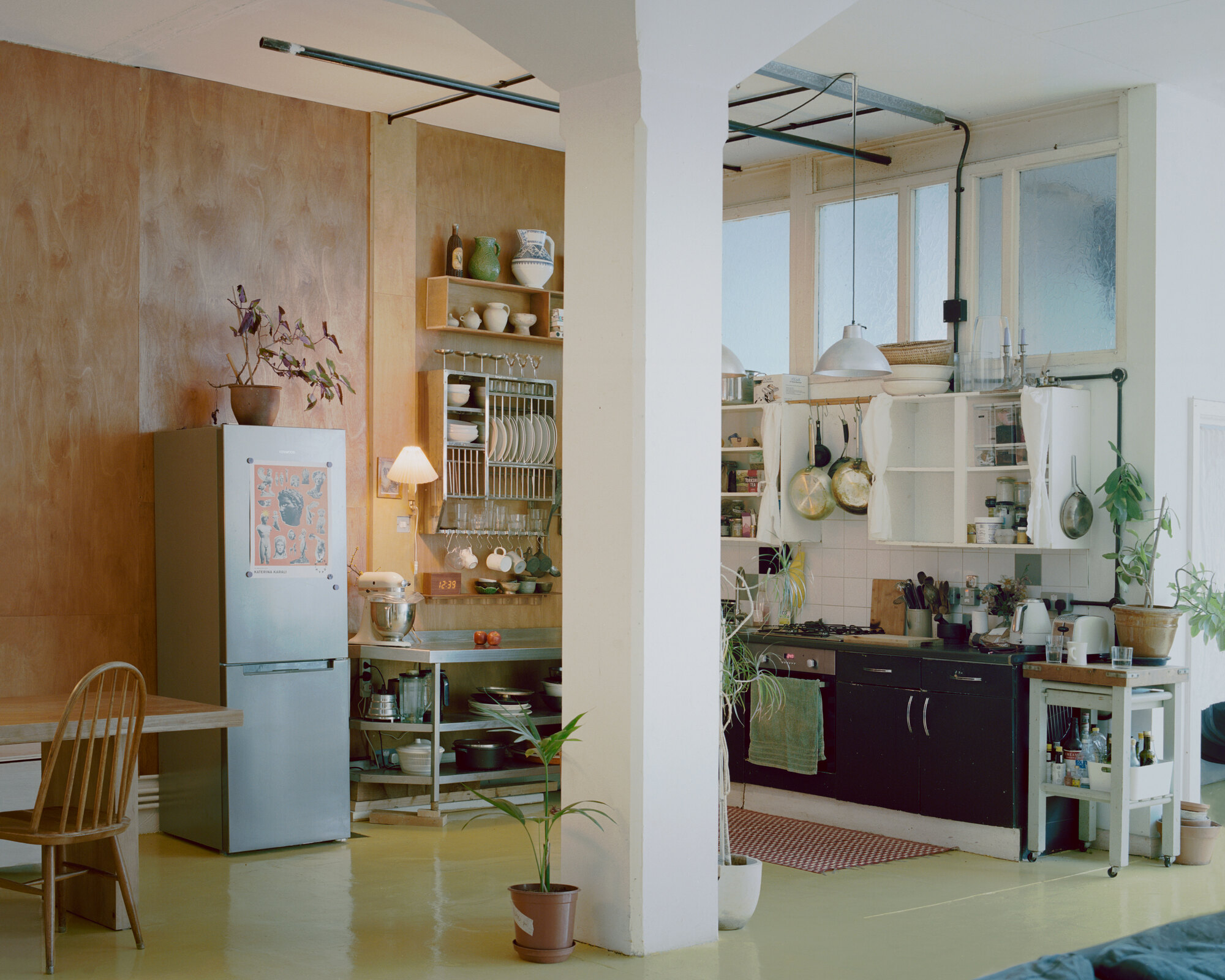 Kitchen alcove with open shelving and dish rack beside plywood partition and exposed piping