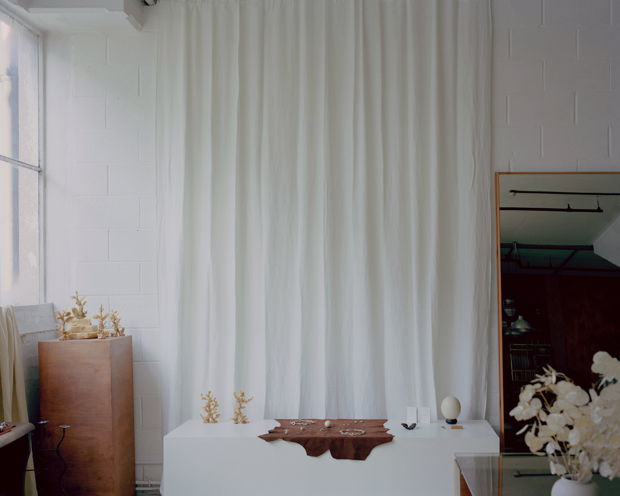 White-painted exhibition room with floor-to-ceiling curtain divider and copper planter beside the window