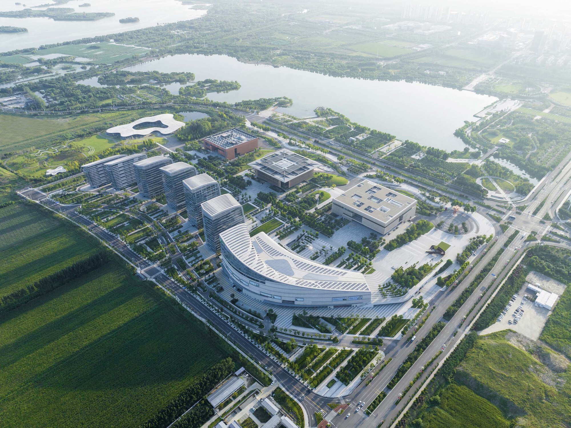 Aerial view of the campus showing curved building volumes and flat-roofed blocks beside green fields and waterways