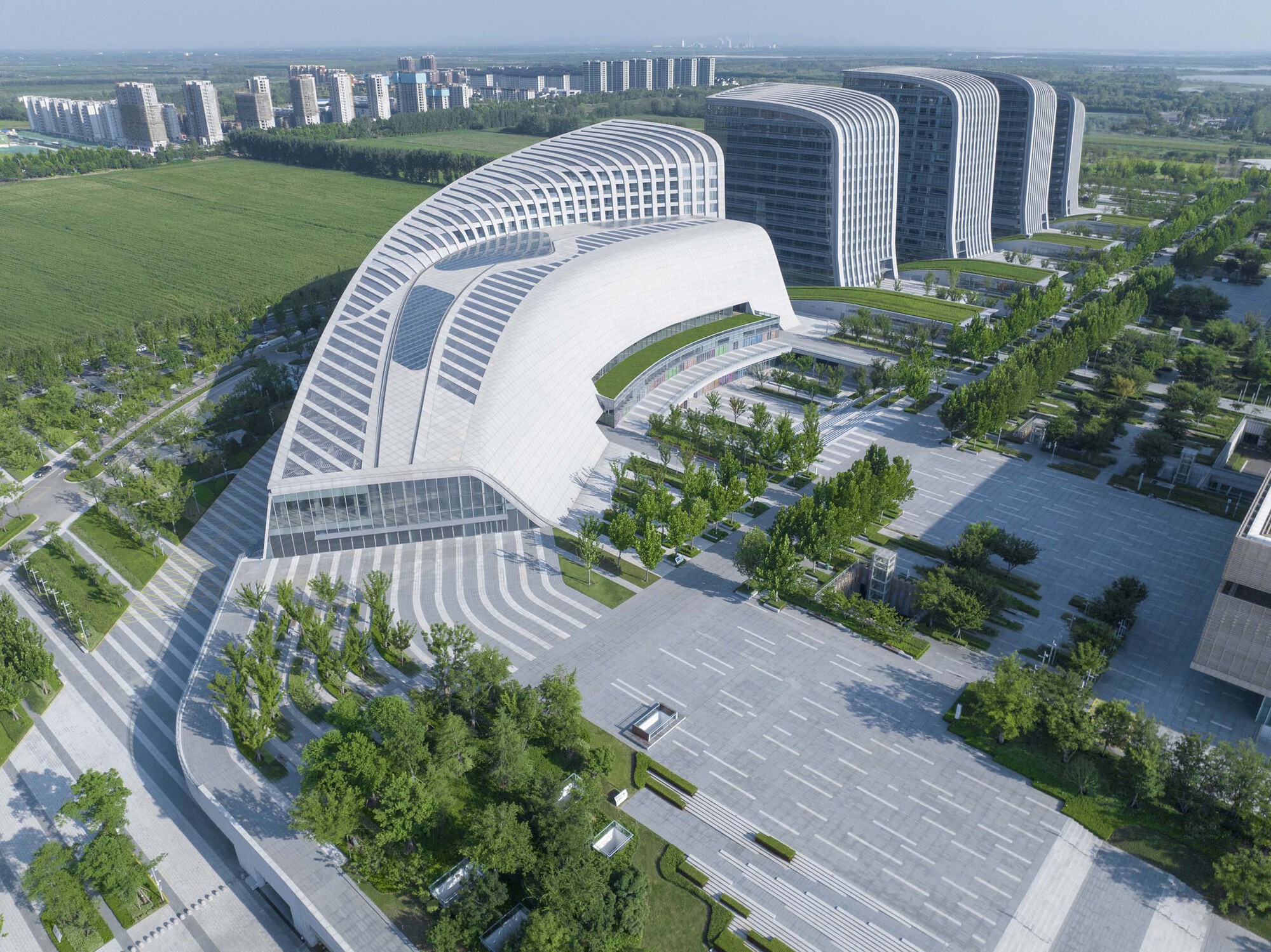 Curved building with horizontal white louvers wrapping a courtyard and entrance plaza under a clear blue sky