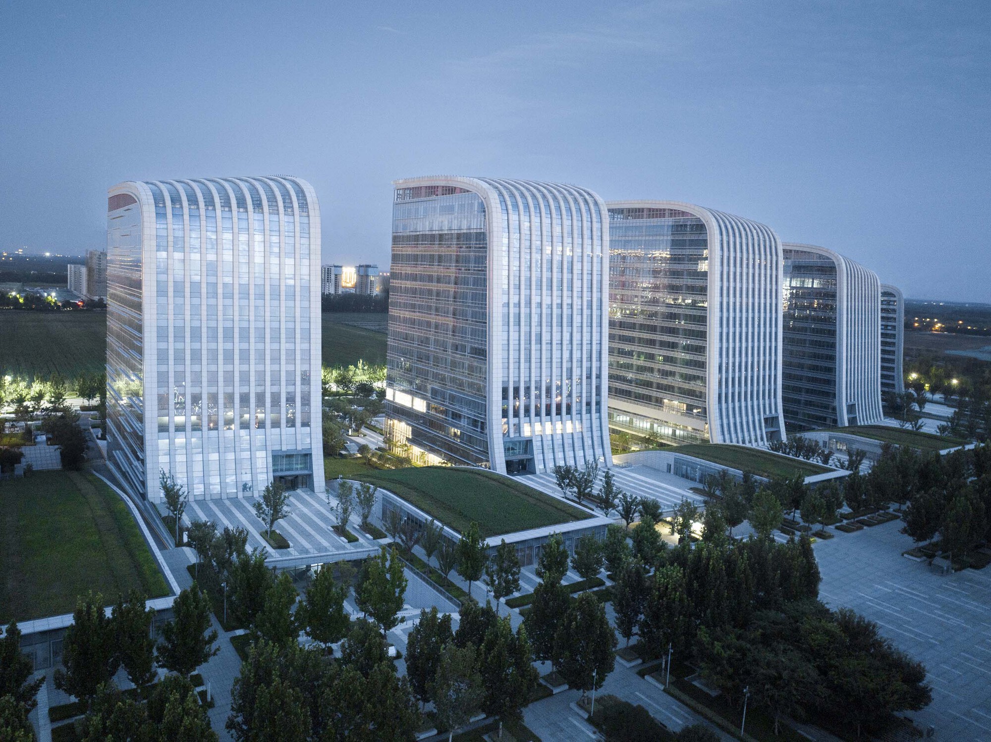 Row of illuminated white-ribbed glass towers at dusk surrounded by landscaped lawns and street trees