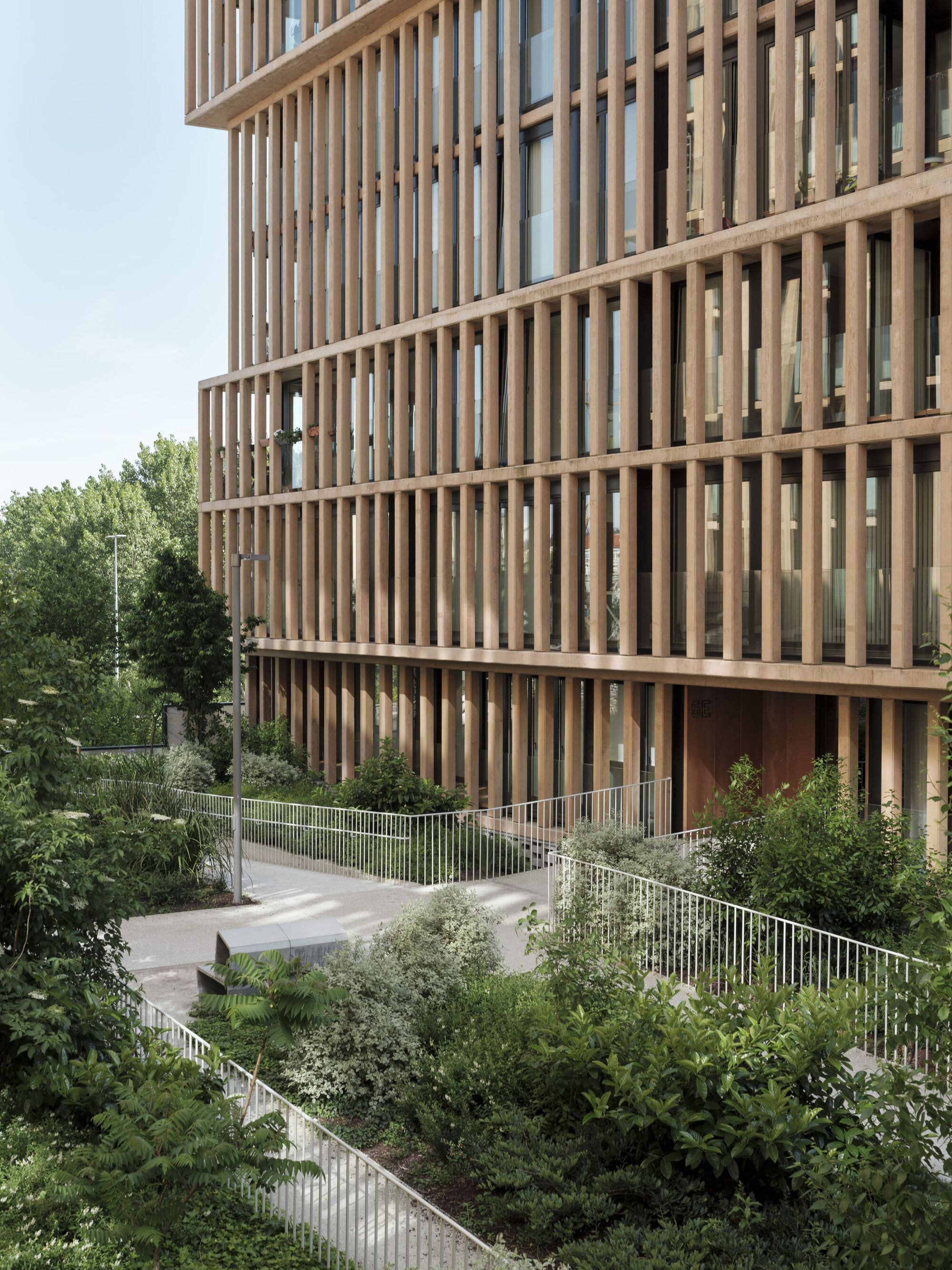Curving pathway with metal railings and planted beds beneath the timber-louvered tower facade
