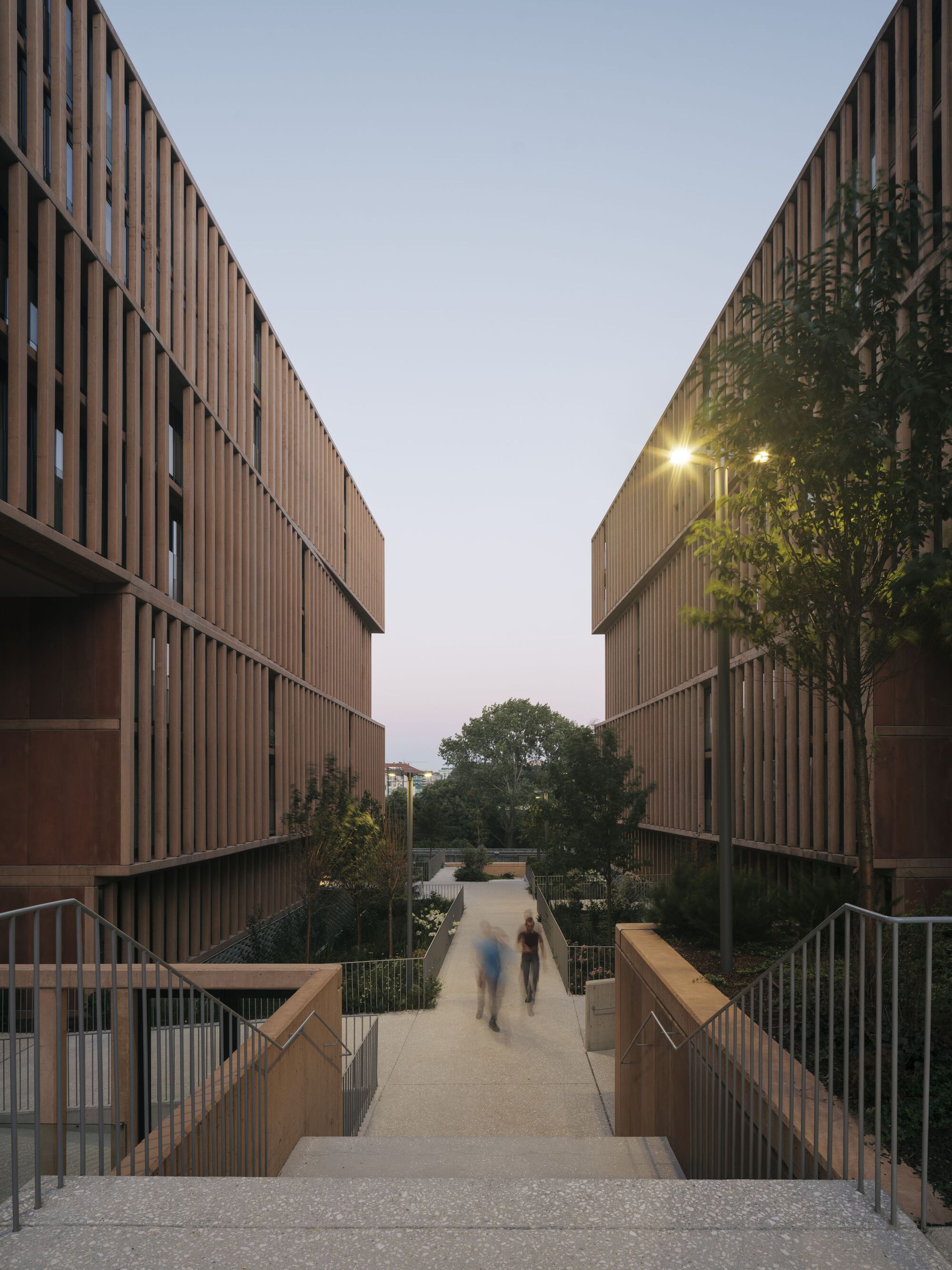 Pedestrian bridge between two timber-clad buildings with residents walking at dusk