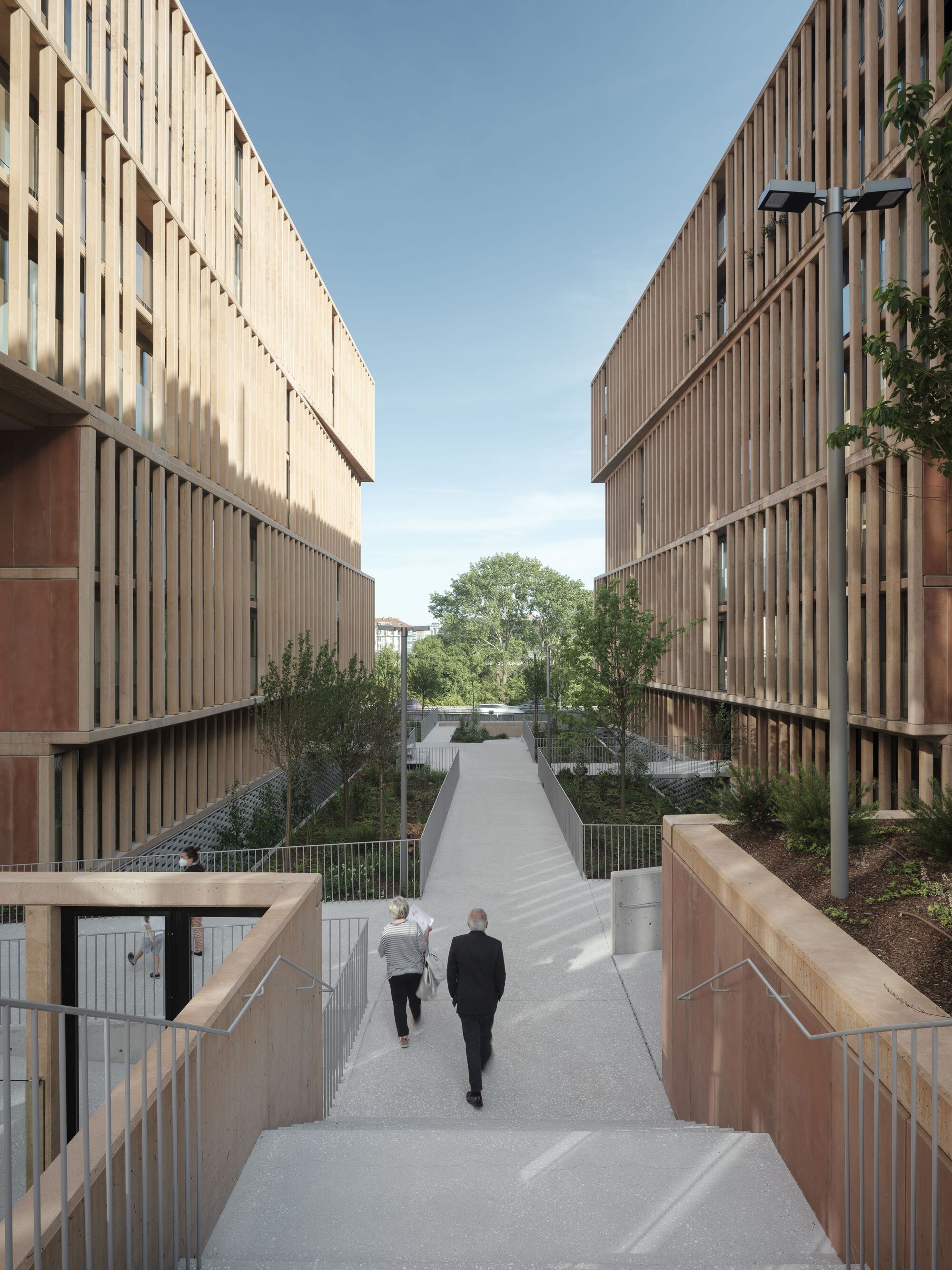 Pedestrian pathway between vertical timber-screened facades with two people descending the sloped walkway