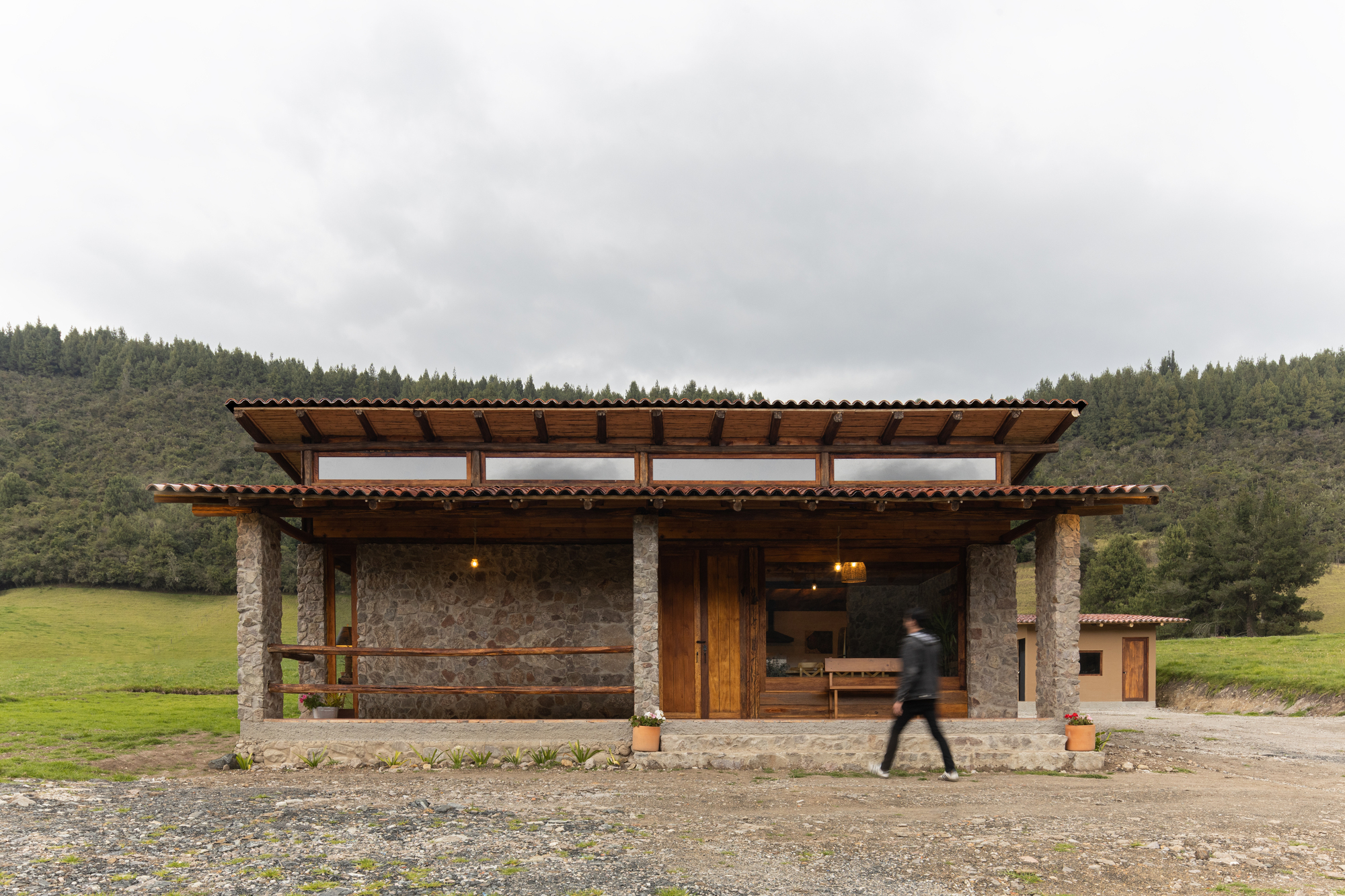 Stone pavilion with timber roof and clerestory windows as a figure walks past in an overcast rural landscape