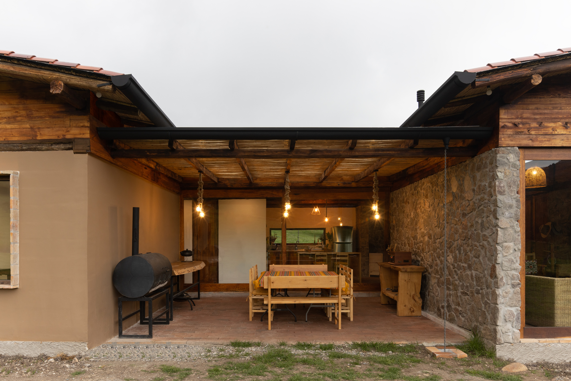 Covered outdoor terrace with exposed timber beams and pendant lights connecting two stone column wings at twilight