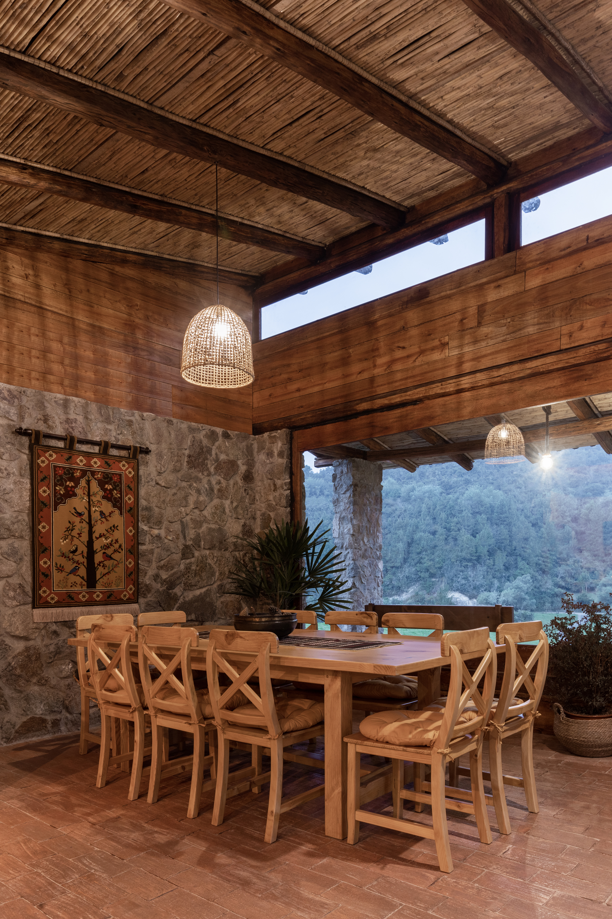 Open dining pavilion with woven pendant lamps and mountain view through timber columns at dusk