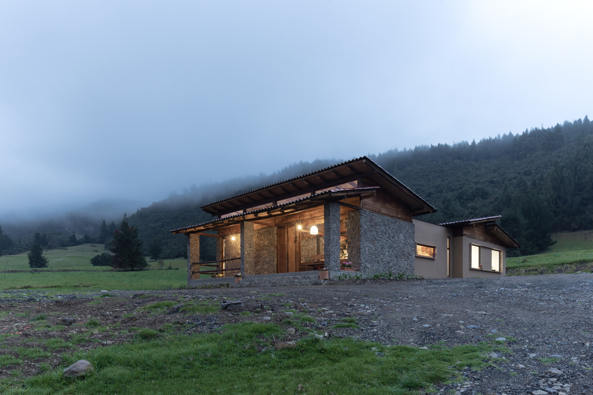 Front facade of the low-slung residence with stone and timber under misty forested hills at twilight