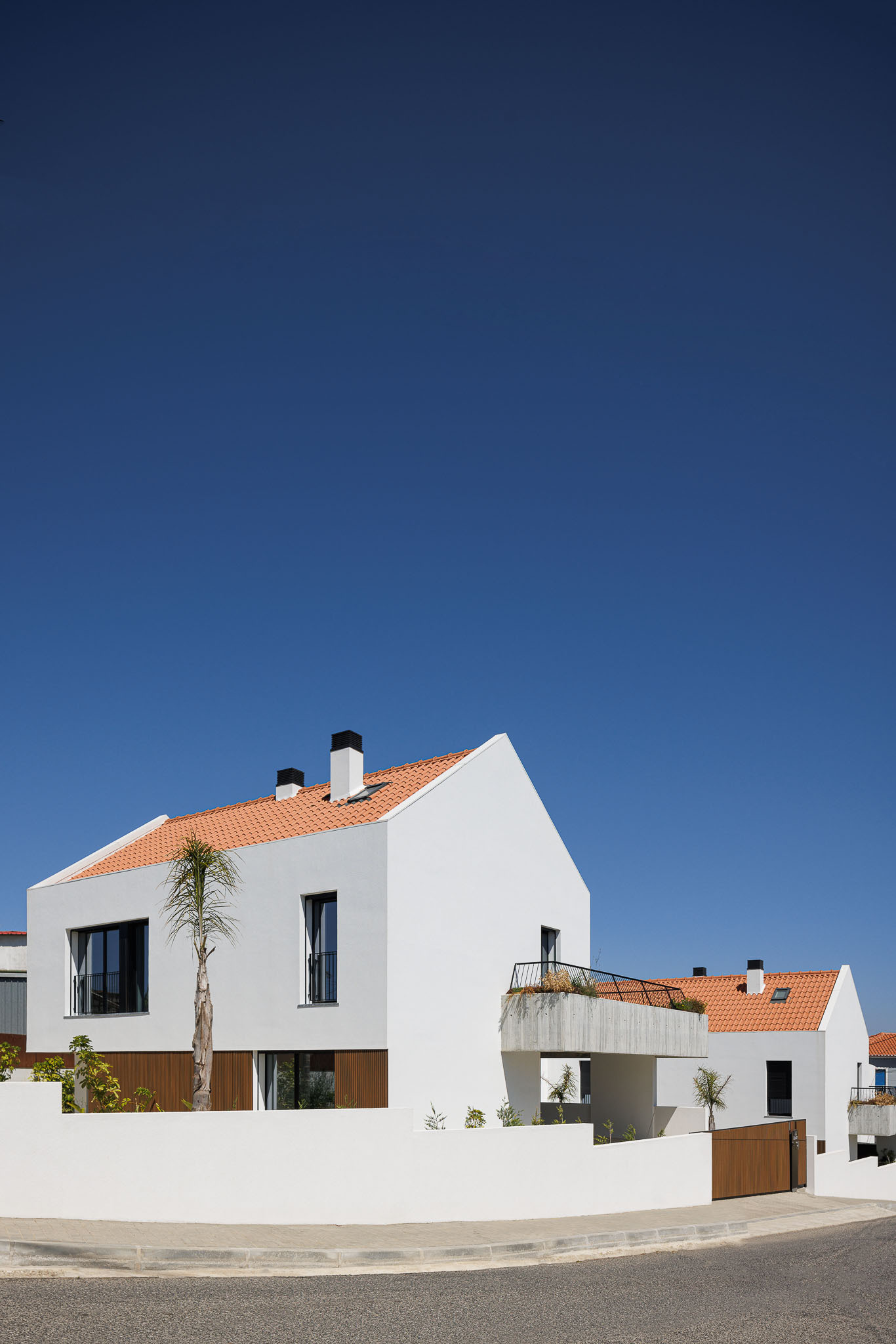 Street view of white plastered volumes with terracotta tile roofs and a palm tree under clear blue sky